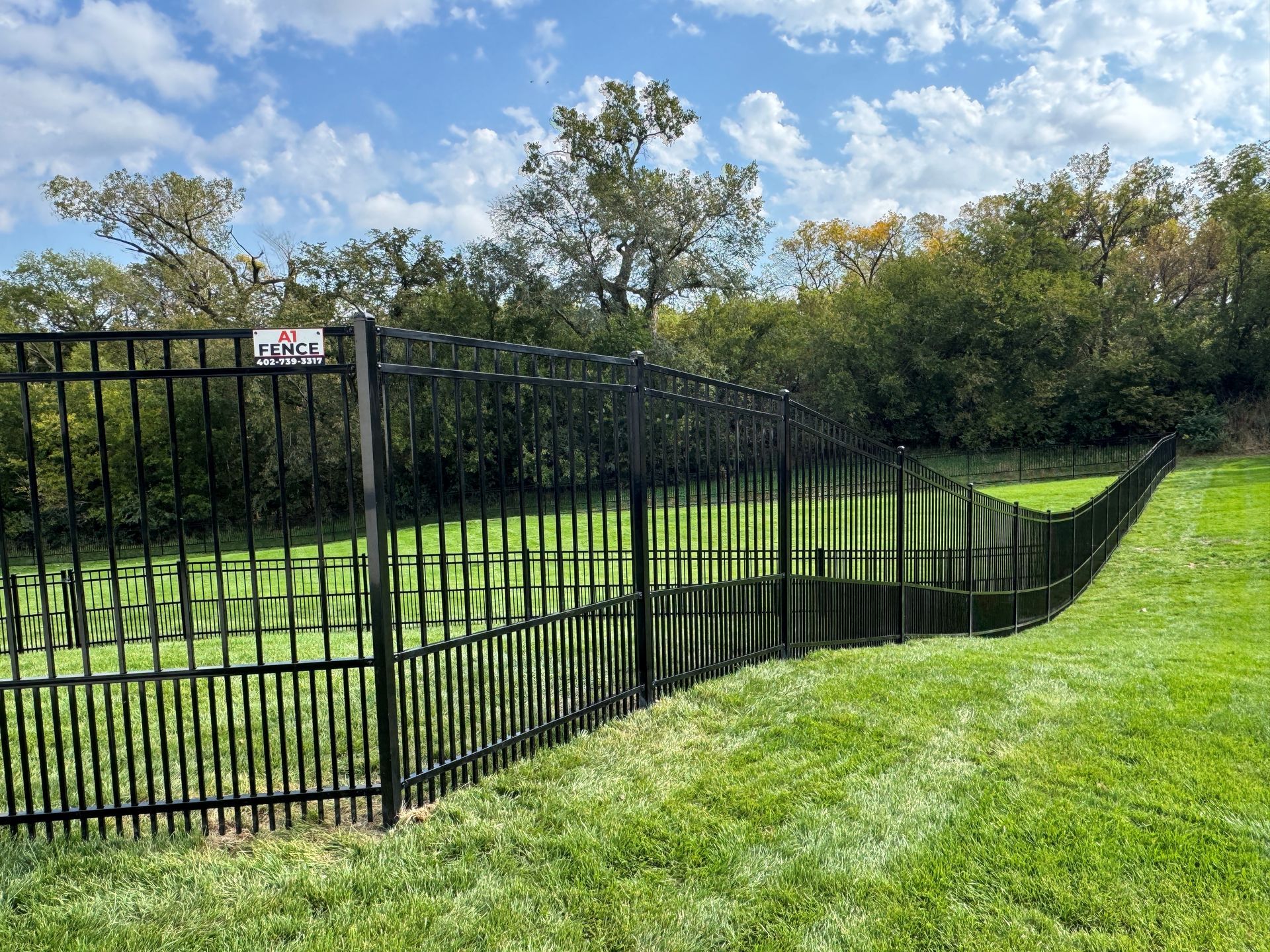 Black metal fence on green grass, curving along a grassy hill with trees in the background under a blue sky.