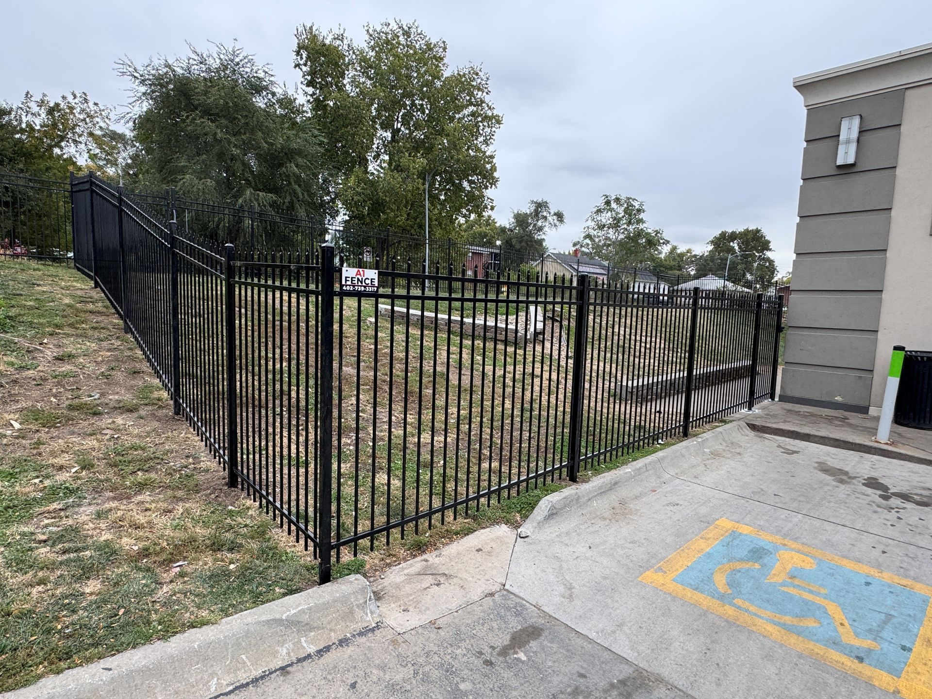 Black metal fence surrounding an area with grass and trees next to a building. Sidewalk with handicap symbol visible.