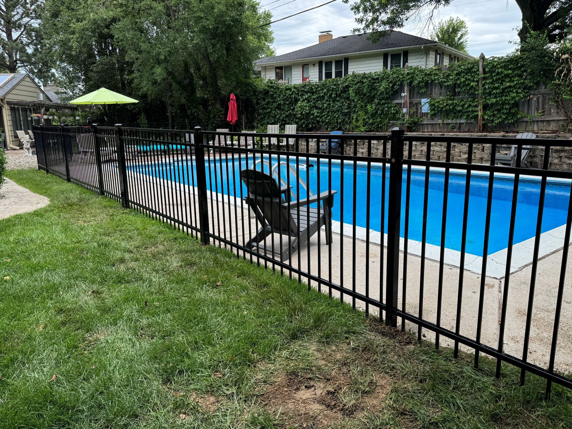 Black metal fence surrounds a blue swimming pool in a backyard. Lawn in foreground, house in background.