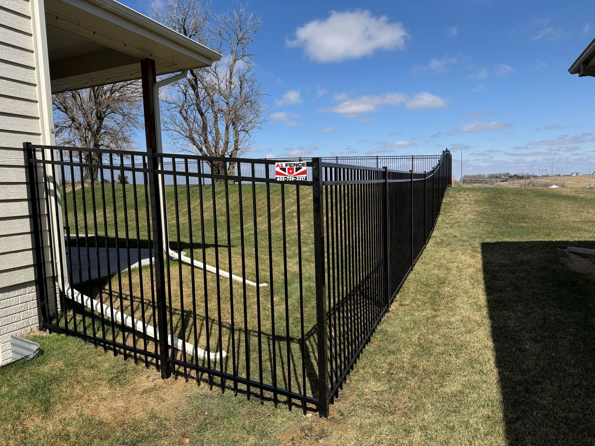 Black metal fence surrounding a yard with grass and a house under a blue sky.