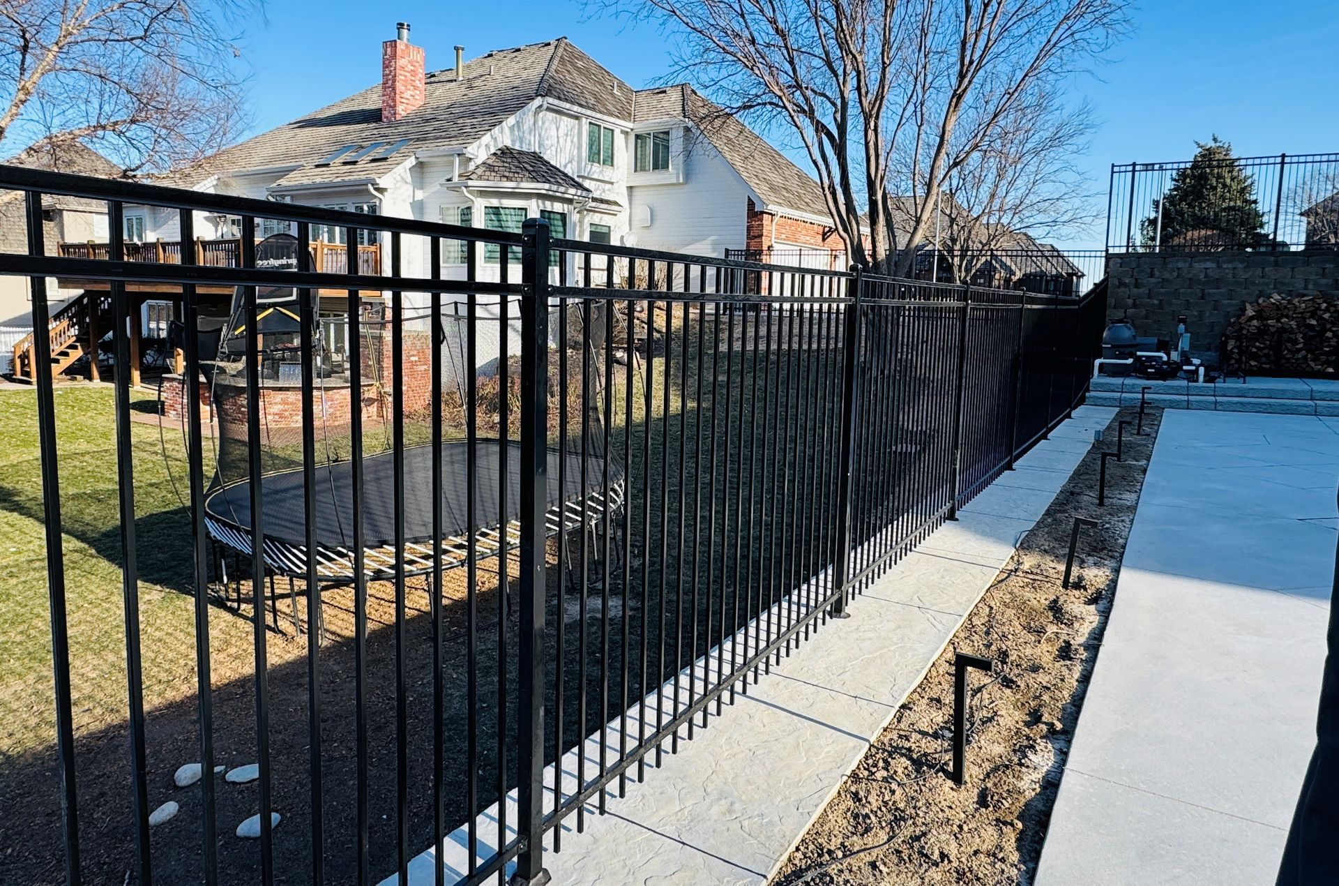 Black metal fence bordering a concrete walkway. Large white house in the background. Sunny outdoor setting.
