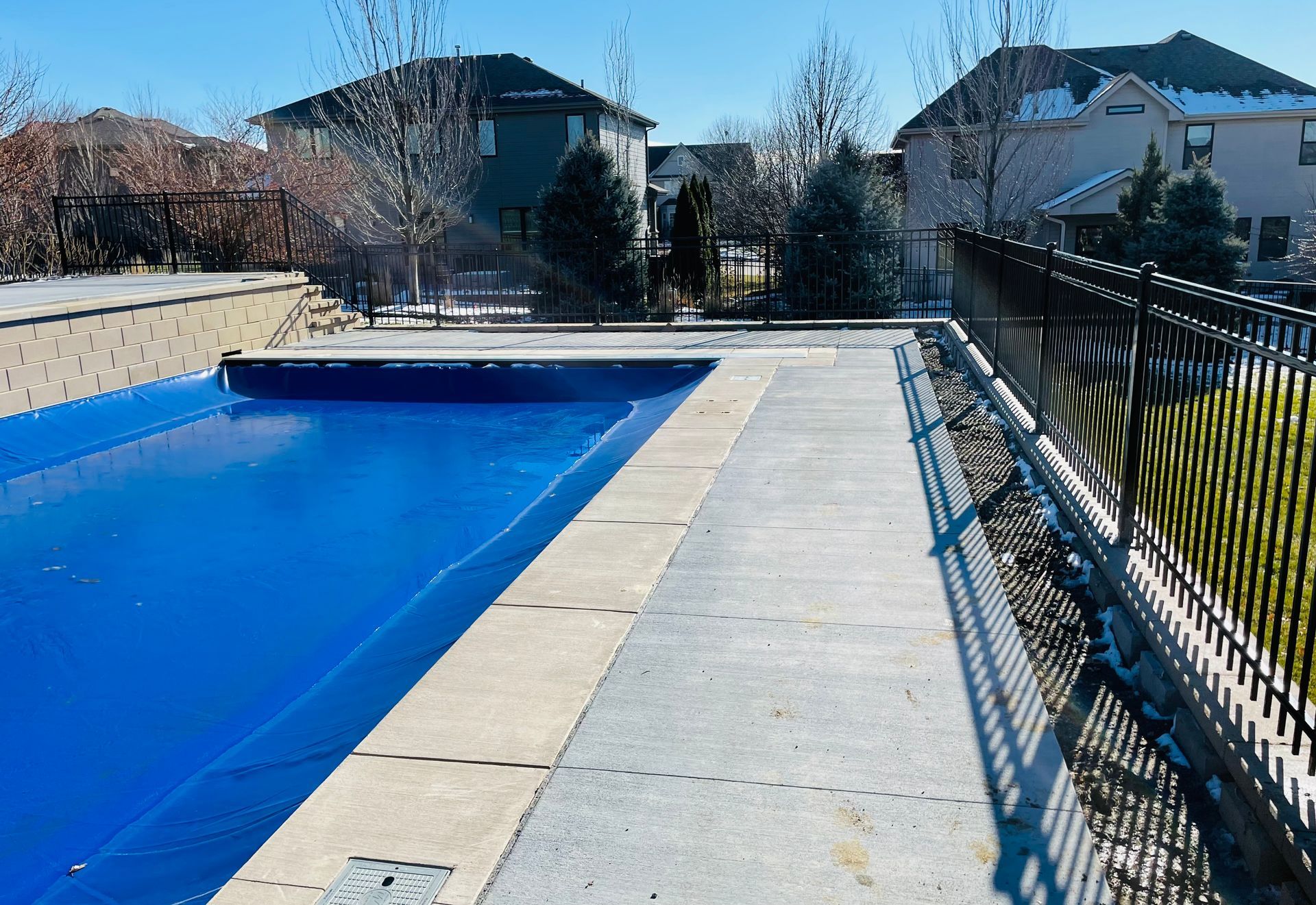 Blue covered pool and concrete patio with black fence on a sunny day. Houses in the background.