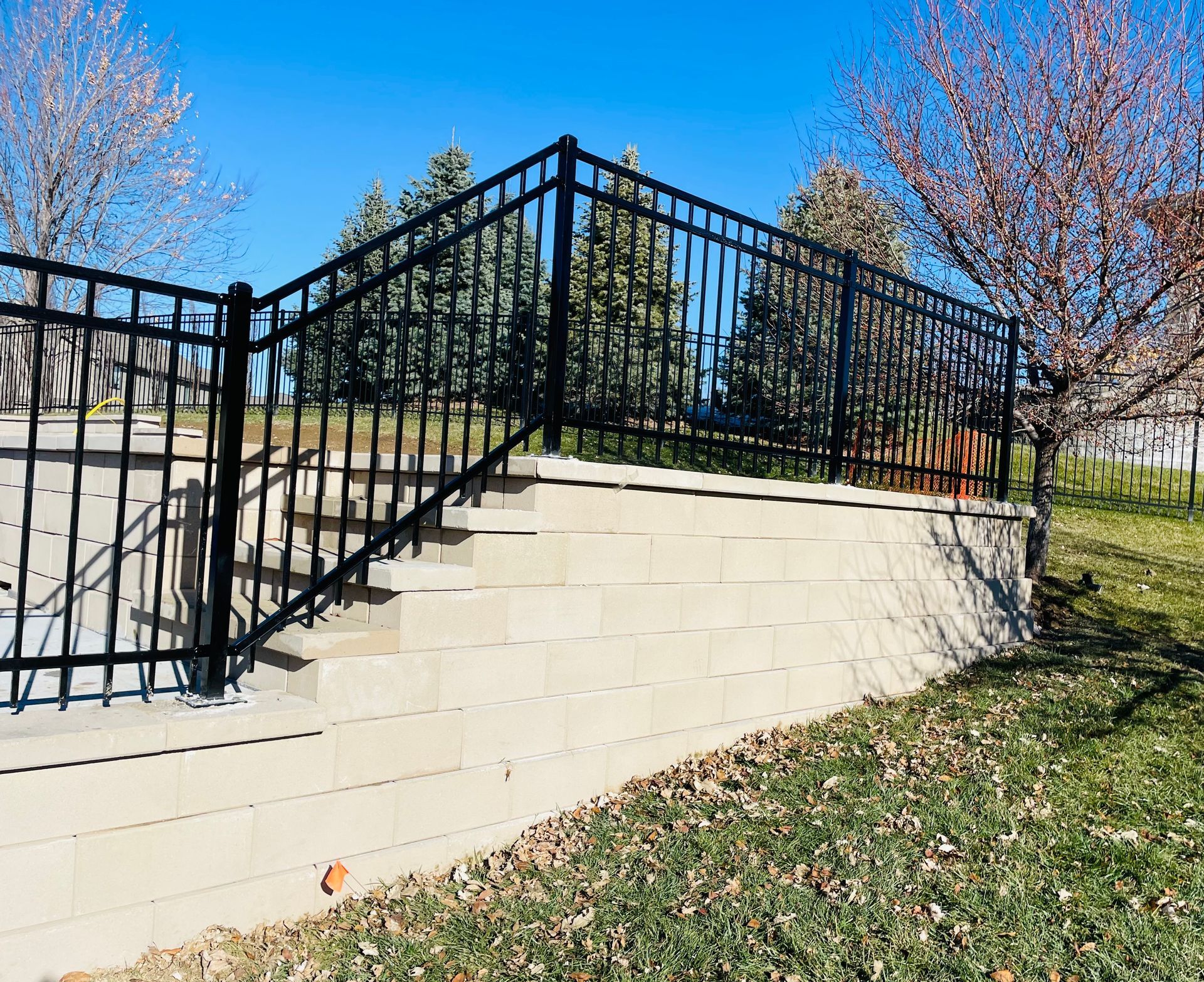 Black metal fence atop a concrete retaining wall with steps. Sunny day, trees in background.