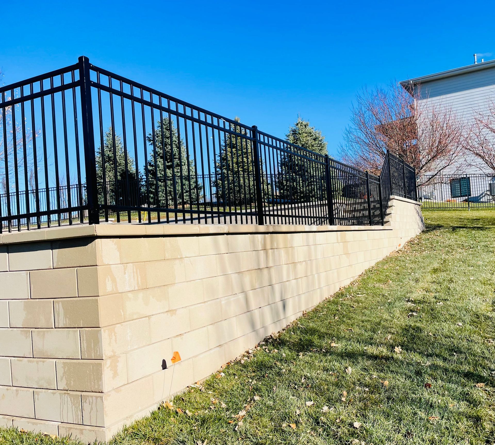 Black metal fence atop a tan brick retaining wall on a grassy slope, with a blue sky background.