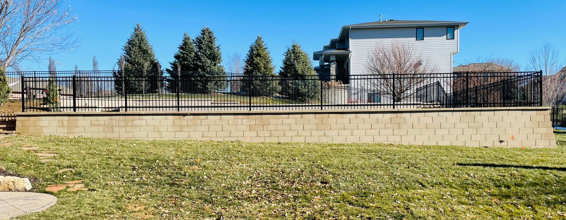 A low retaining wall topped with a black metal fence, behind a grassy yard, with a building in the background.