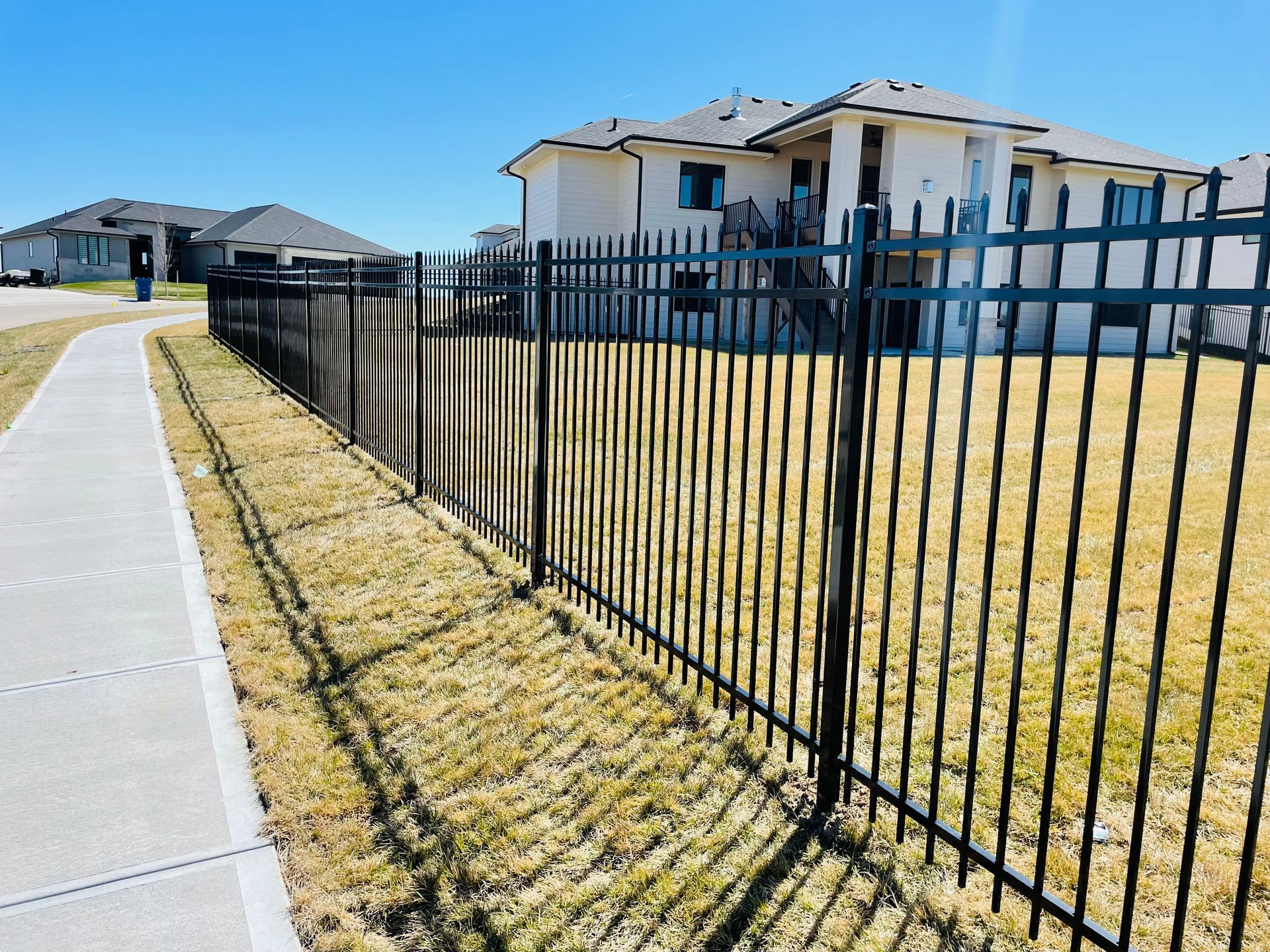 Black metal fence along a sidewalk, bordering a residential lawn. Houses in background under a blue sky.
