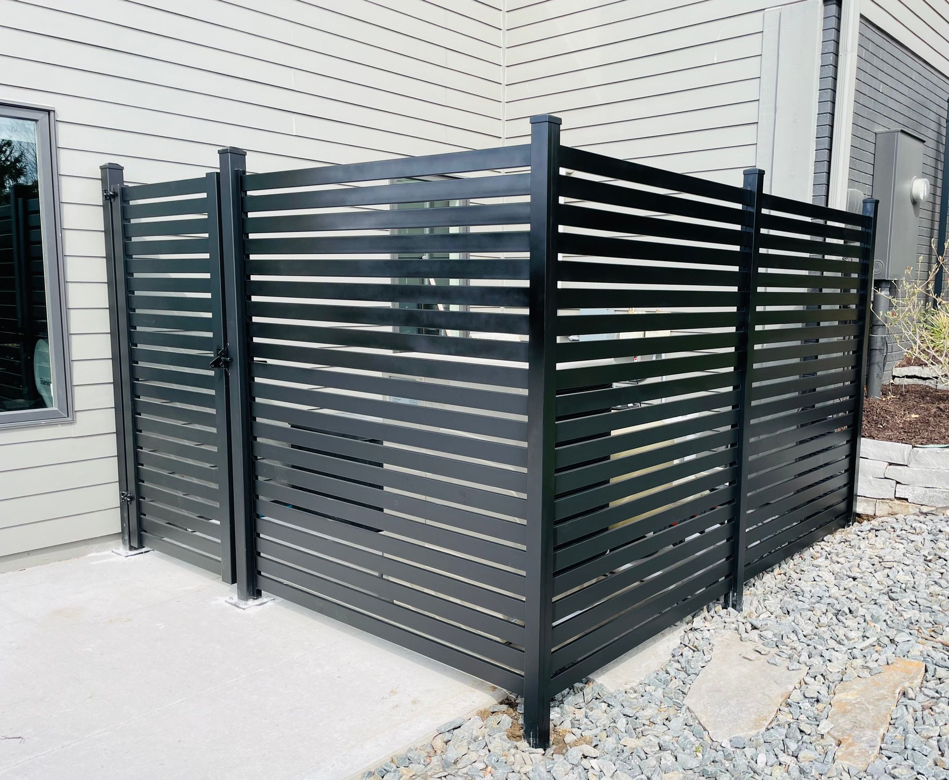 Black slatted privacy fence encloses utility equipment on a concrete patio, adjacent to a house with light siding.