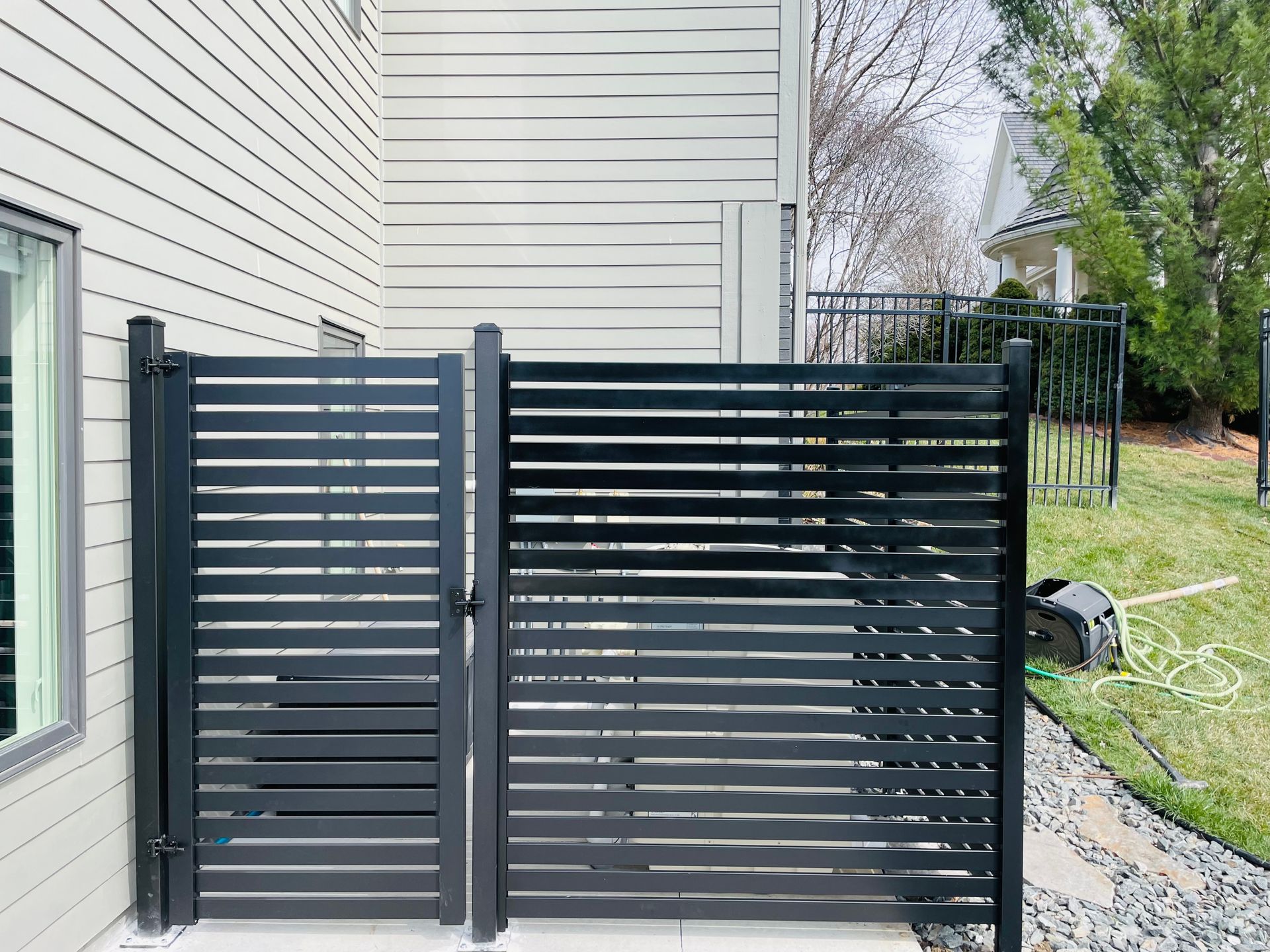 Black horizontal slat gate and fence adjacent to a building. Green grass and trees in the background.
