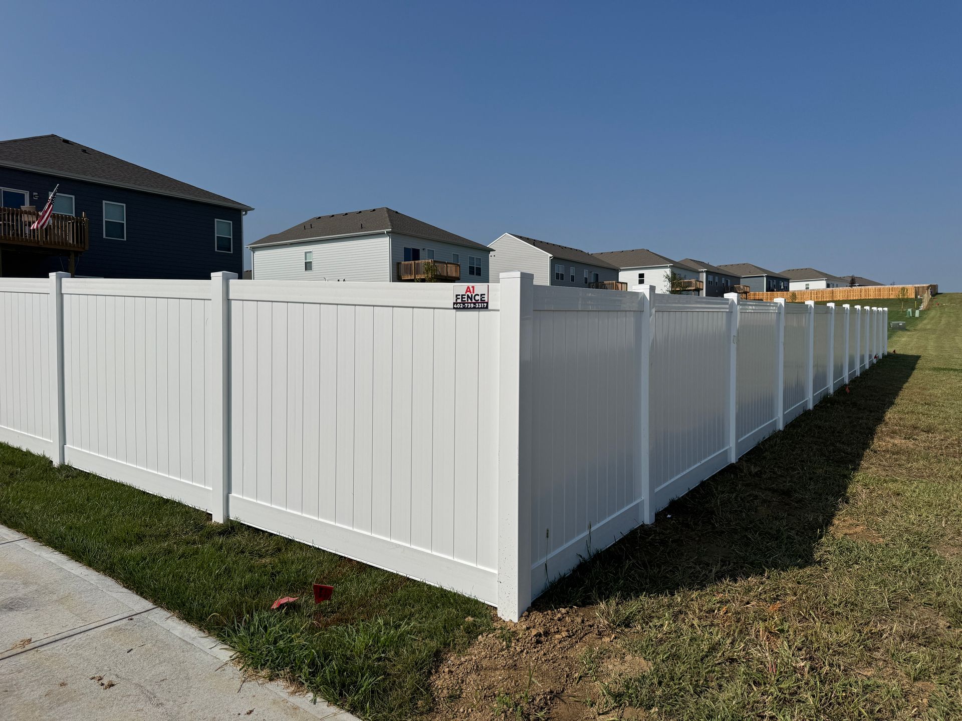 White vinyl fence in front of several houses with green grass and a blue sky.