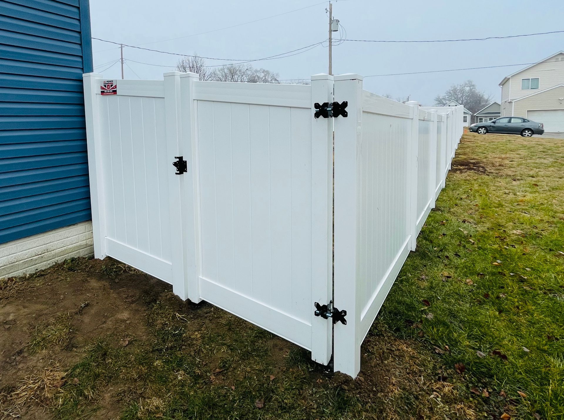 White vinyl fence with gate, bordering a blue house and grassy lawn. Black hinges and latch.