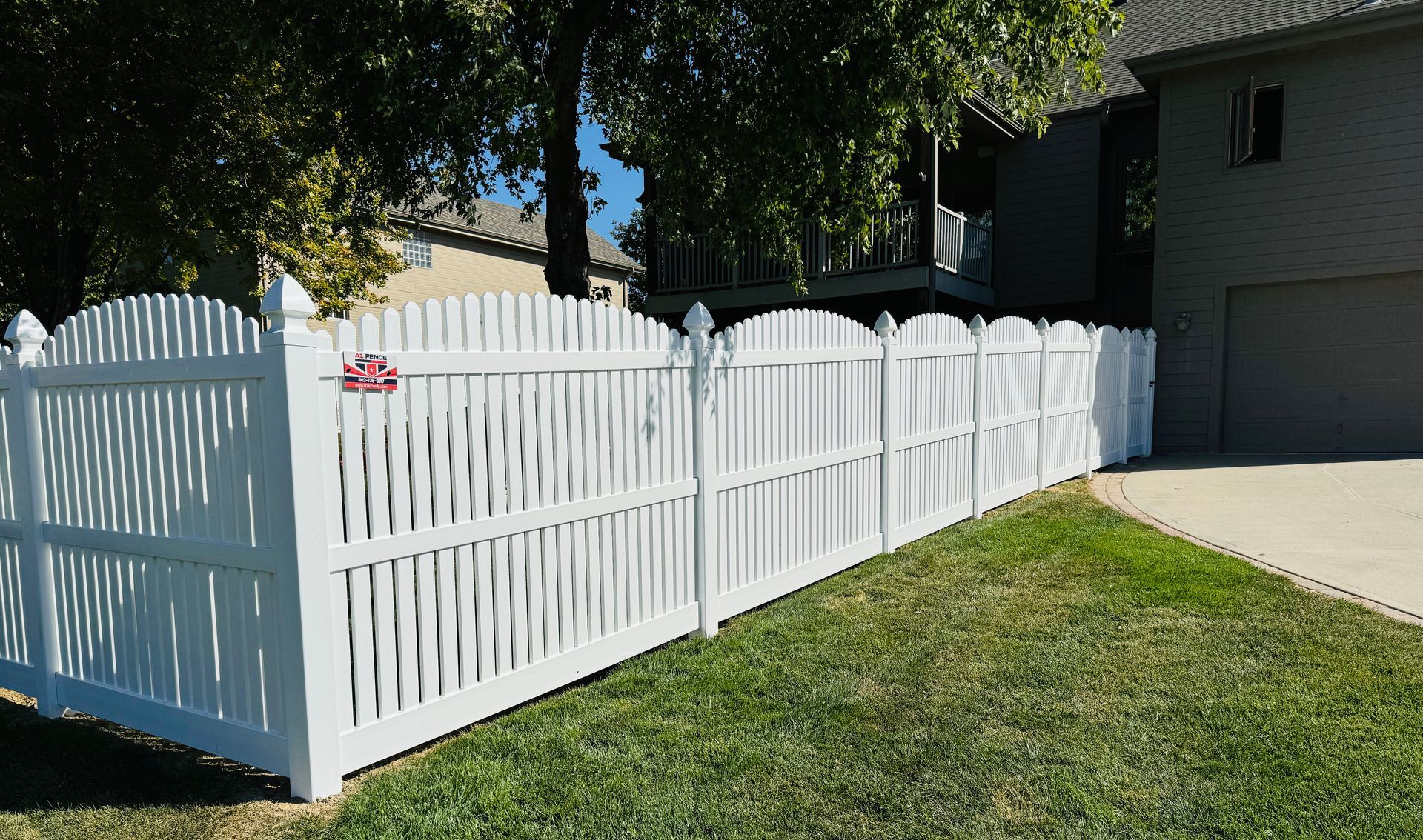 White picket fence encloses a grassy yard; a house and trees are in the background on a sunny day.