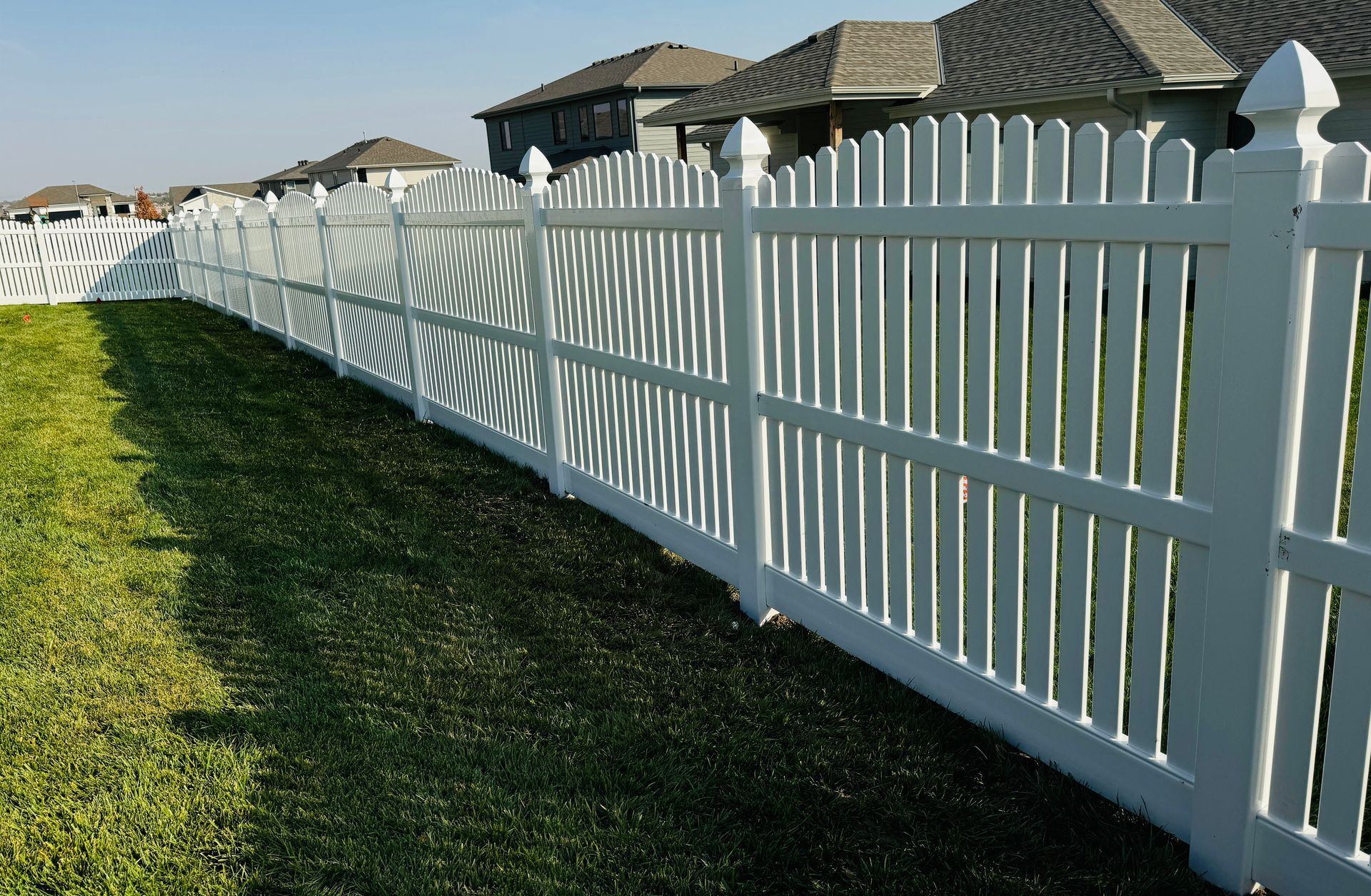 White picket fence in a grassy yard, with houses in the background on a sunny day.