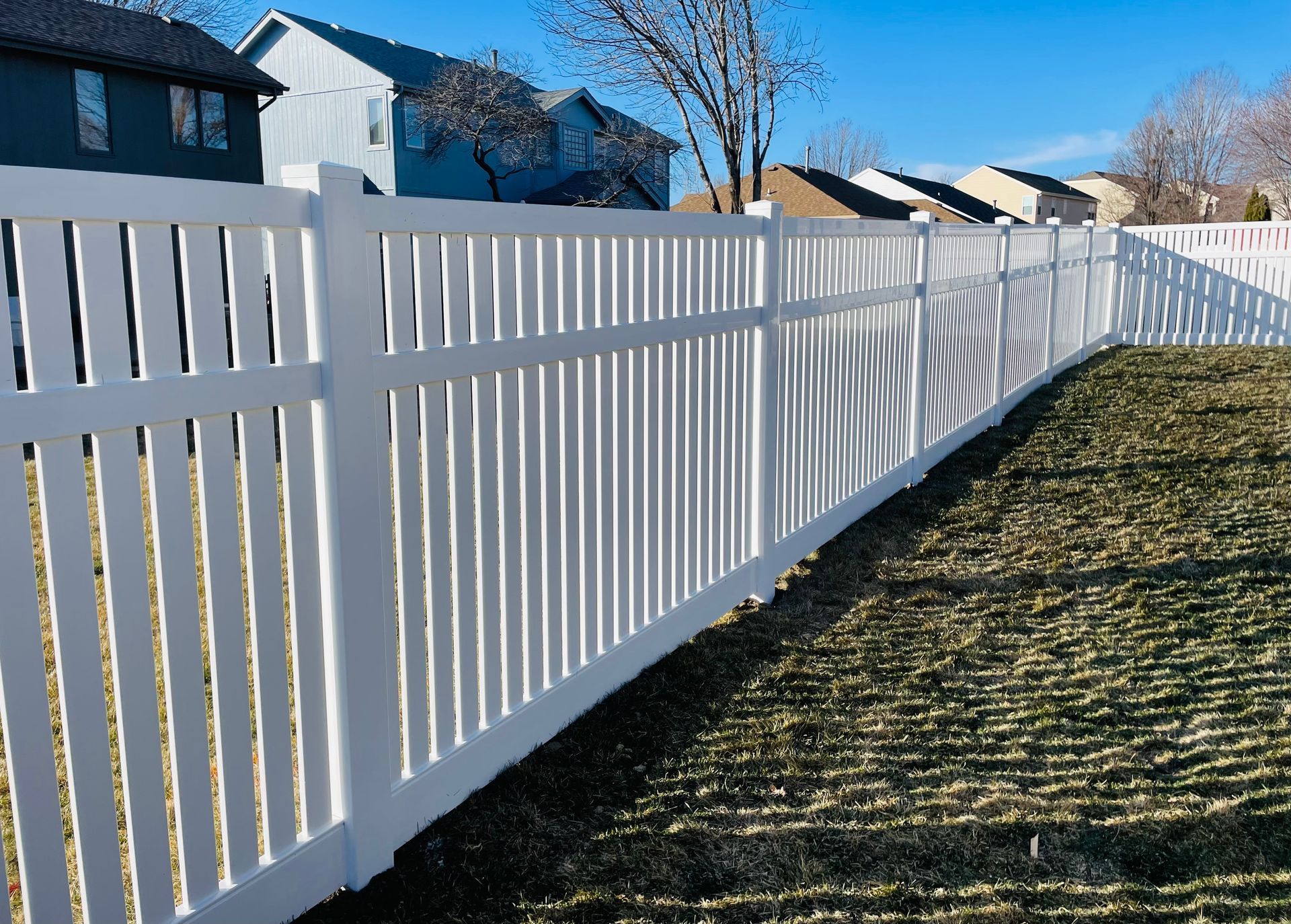 White vinyl fence in a grassy yard, sunlight casts shadows. Houses in background.