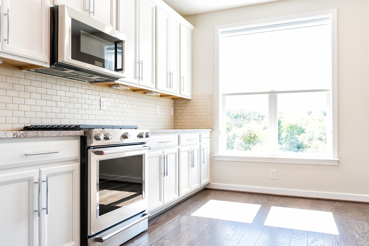 An empty kitchen with white cabinets , a stove , microwave , and a window.