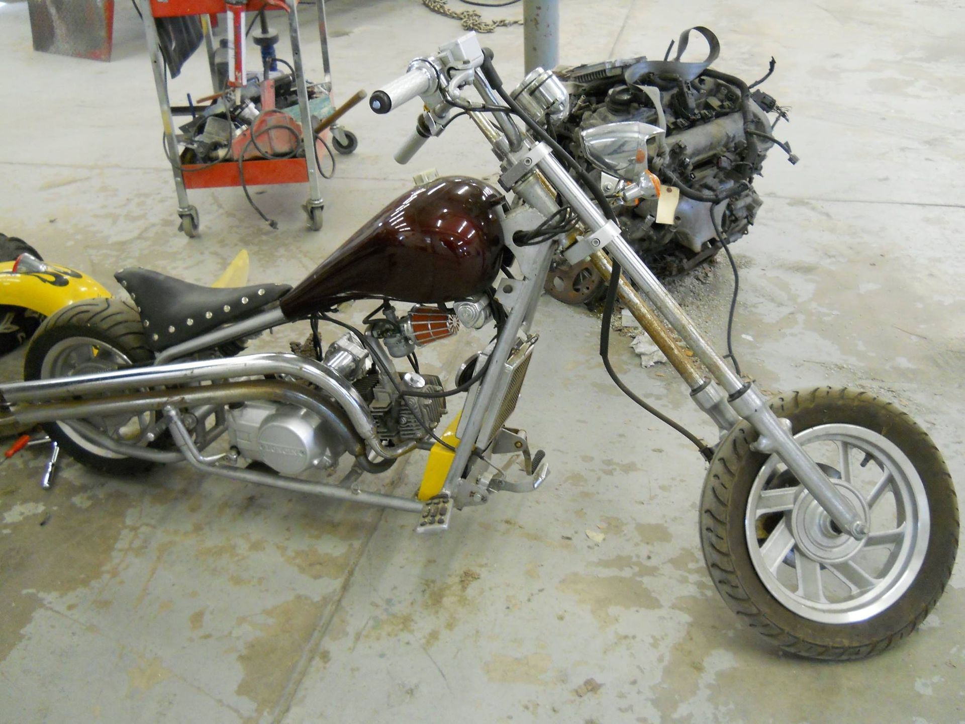 Custom motorcycle frame with maroon tank and chrome exhaust in a workshop, partially disassembled.