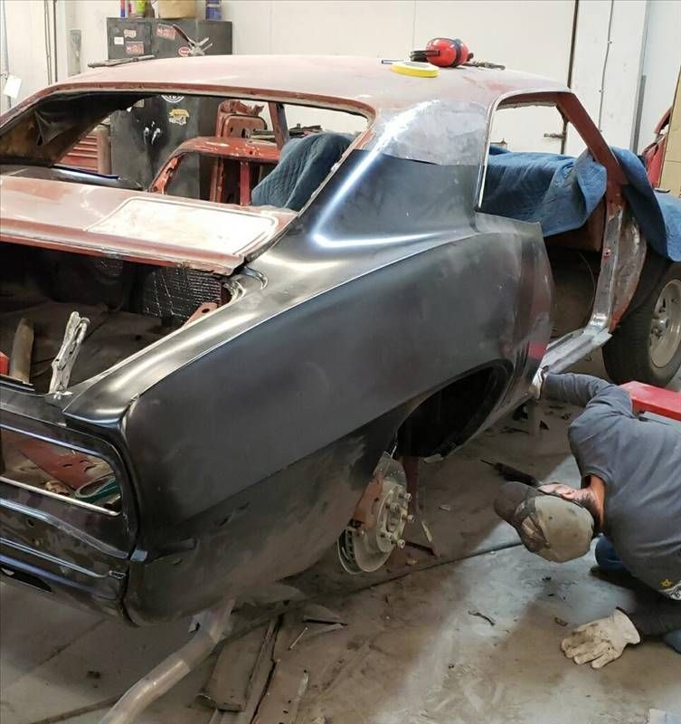 Partially restored classic car in a garage, with a worker sanding the rear quarter panel.