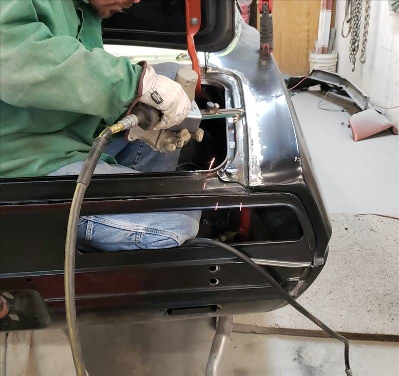 Worker welding a glossy black car body in a garage, using a torch and hose.