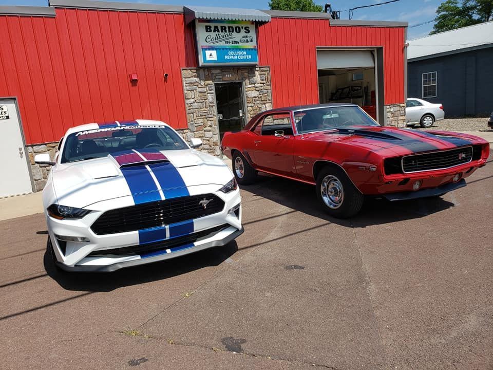 Two classic muscle cars parked outside a red building, one white with blue stripes and one red.