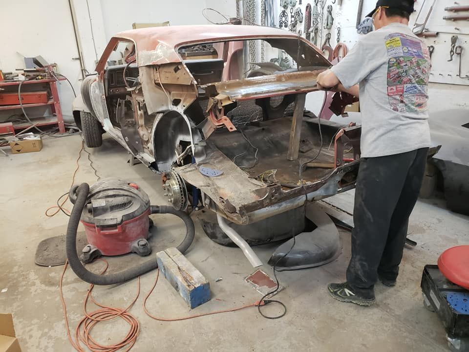 Man working on a stripped car body in a garage, with tools and equipment scattered around.
