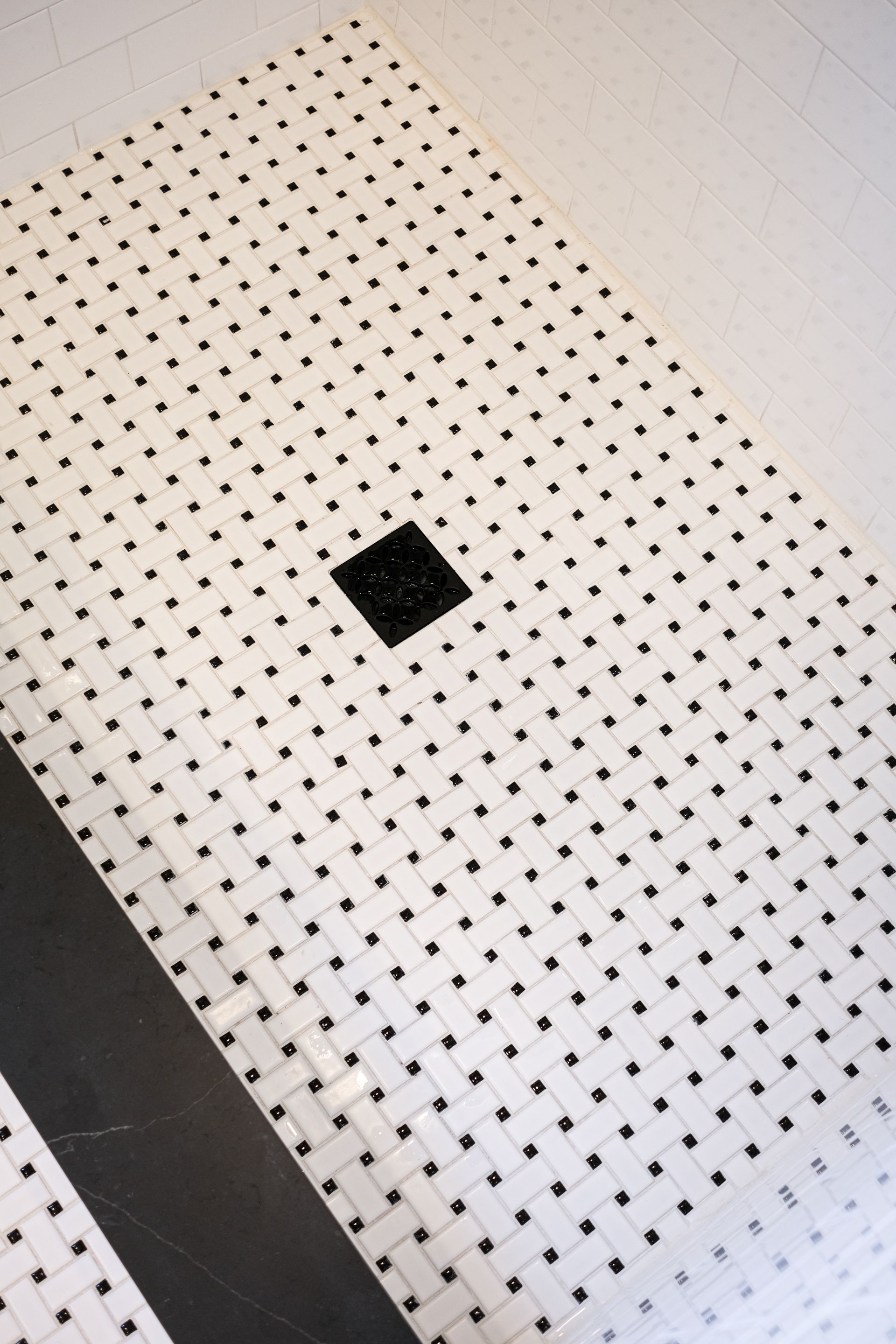 A black square shower drain centered in a white, basket-weave patterned tiled shower floor.