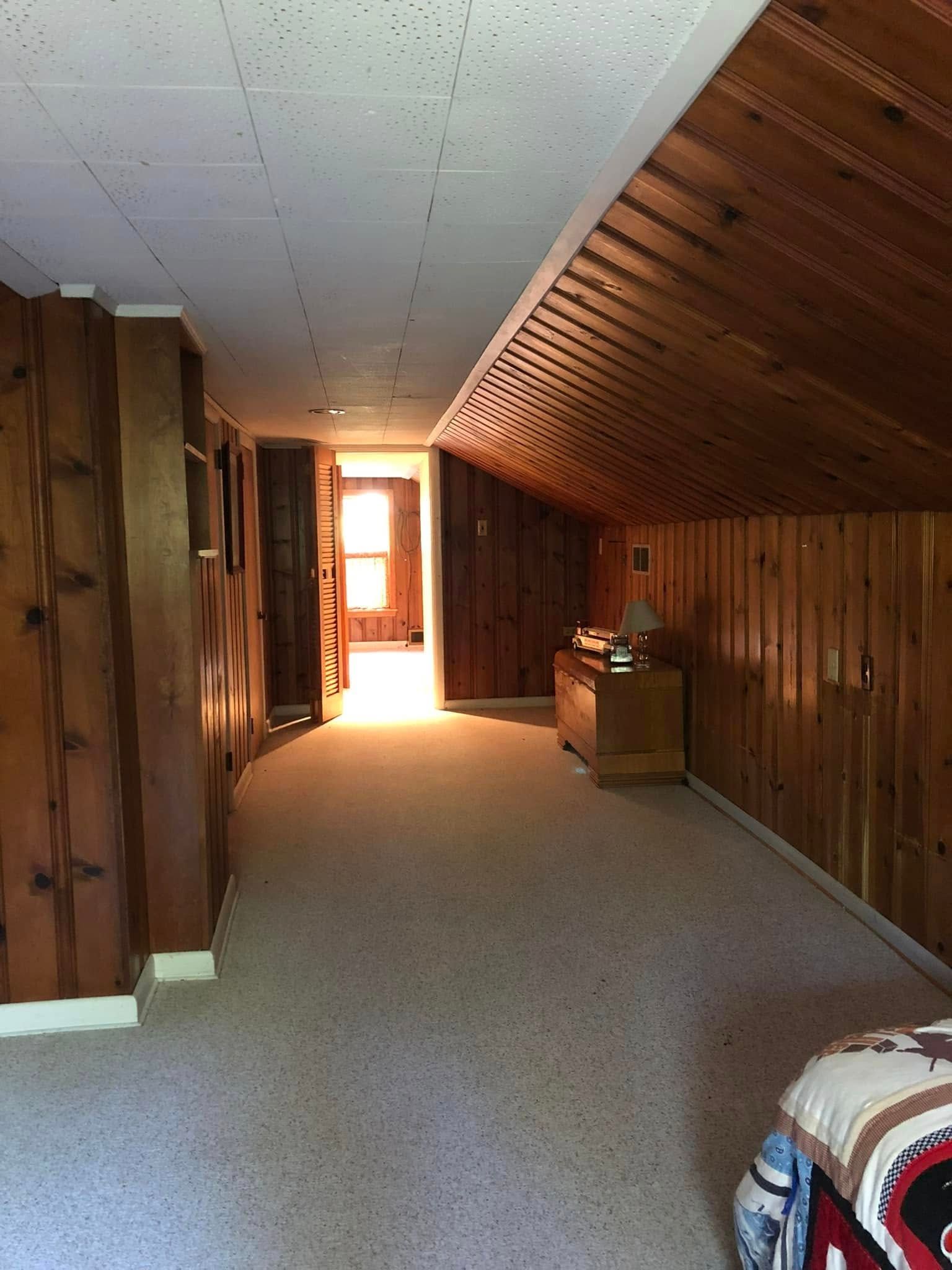 Room with wood paneling on walls and ceiling, carpeted floor, sunlight from doorway at the end of the hall.