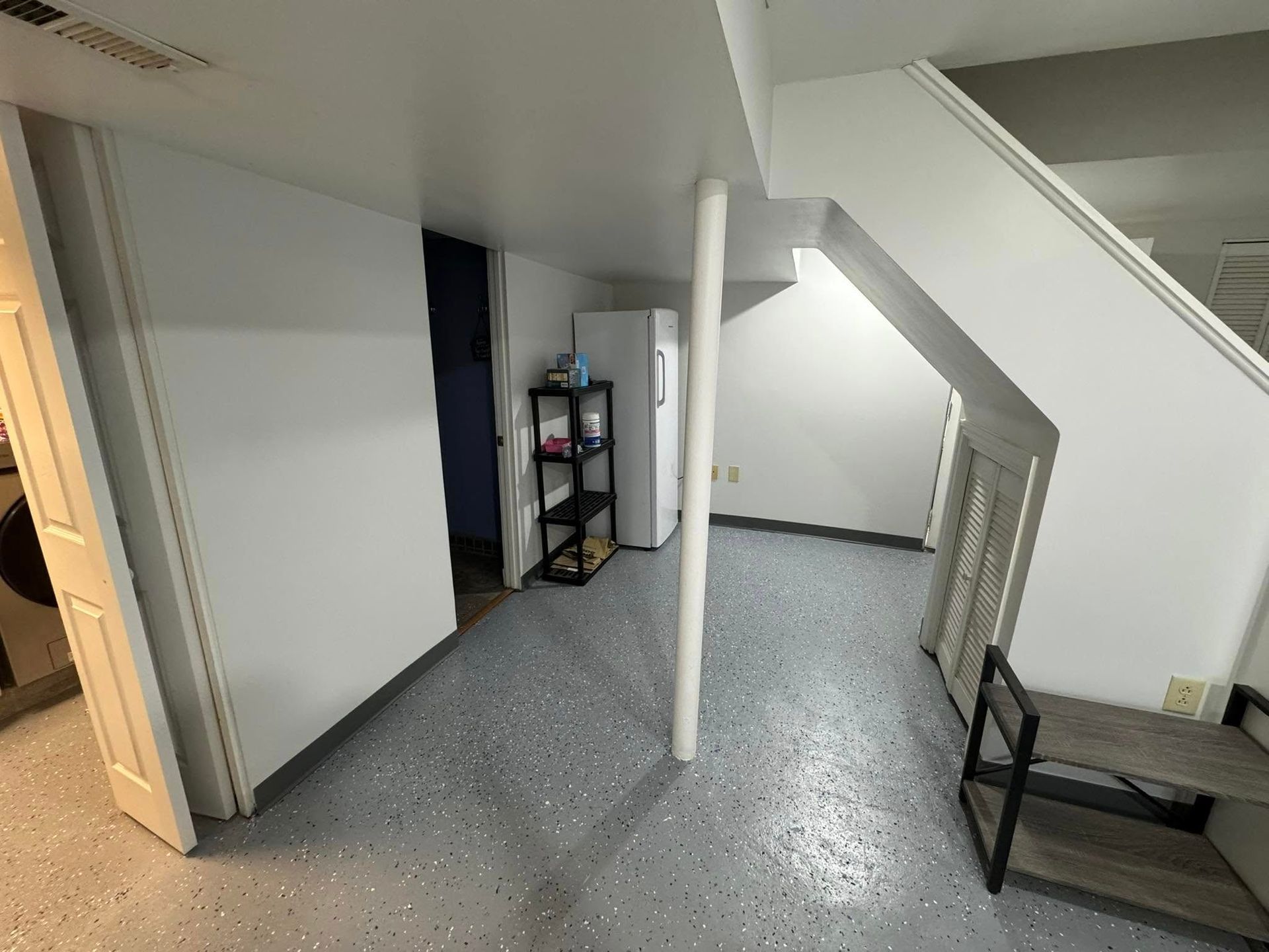 Basement interior: white walls, epoxy floor, staircase, support beam, refrigerator, shelving, and a door.