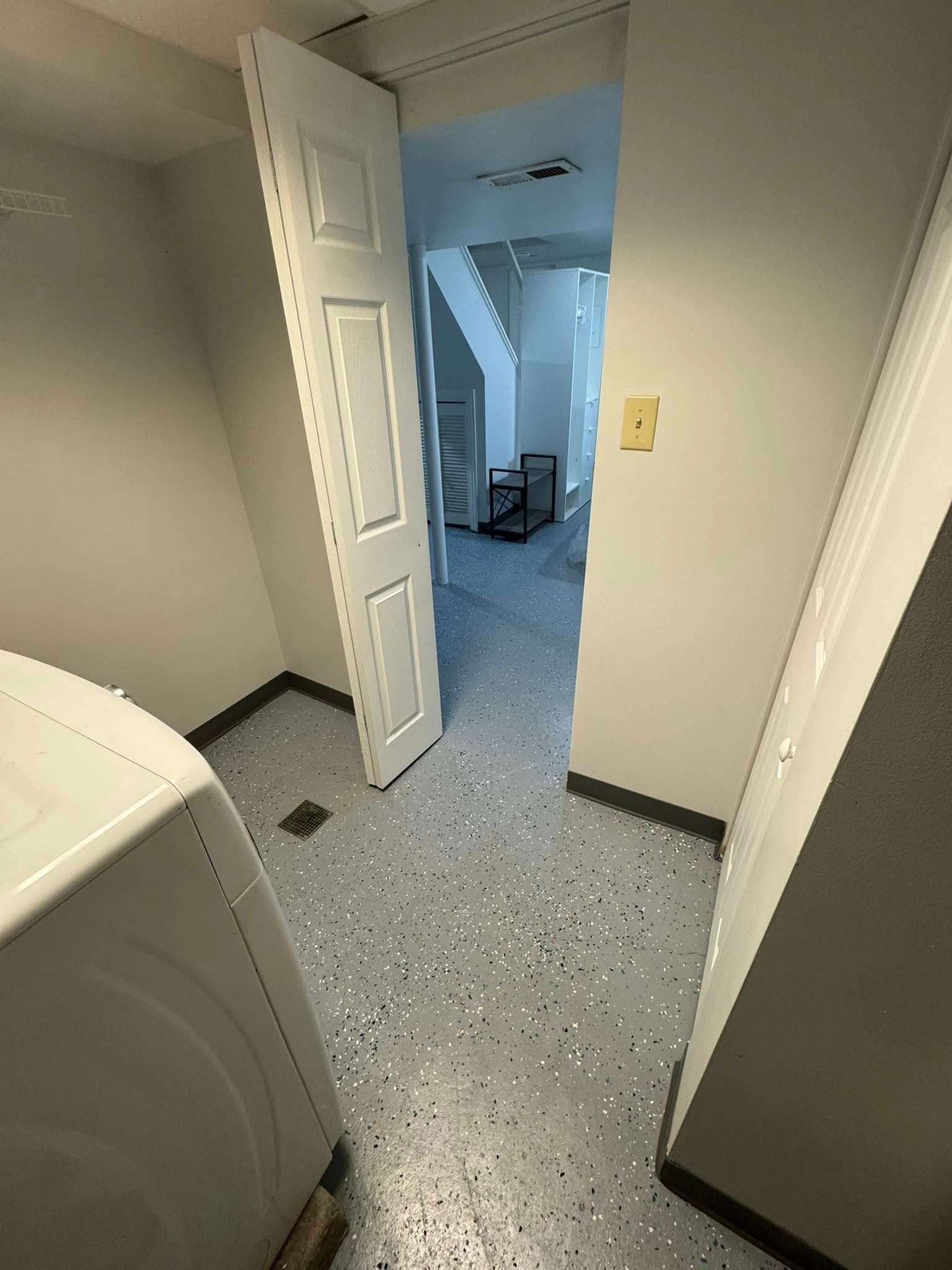 Laundry room with open door leading to hallway with stairs. White appliances, gray speckled floor, light walls.