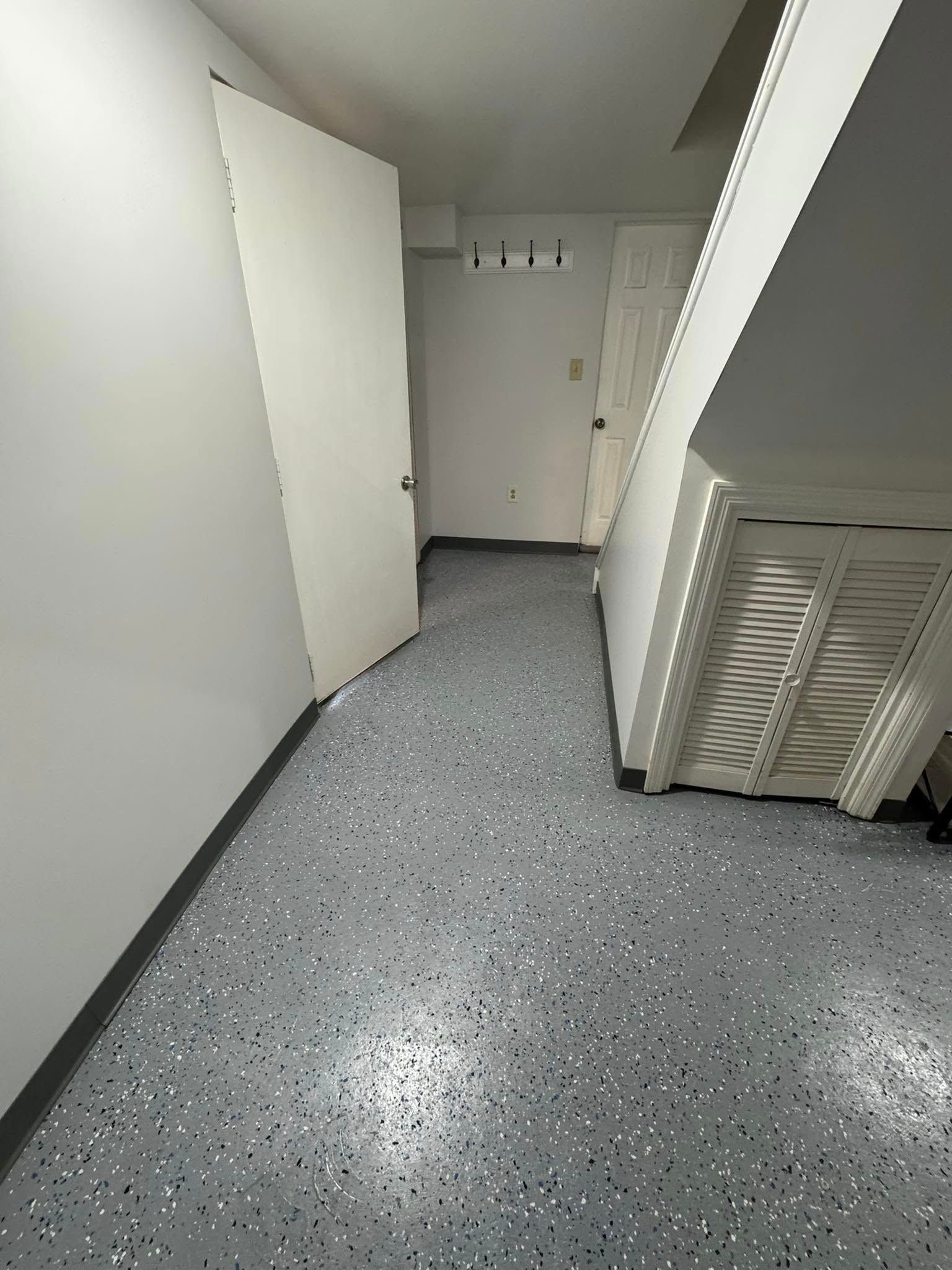 A small, white-walled room with a speckled gray floor. An open white door leads to a further hallway.