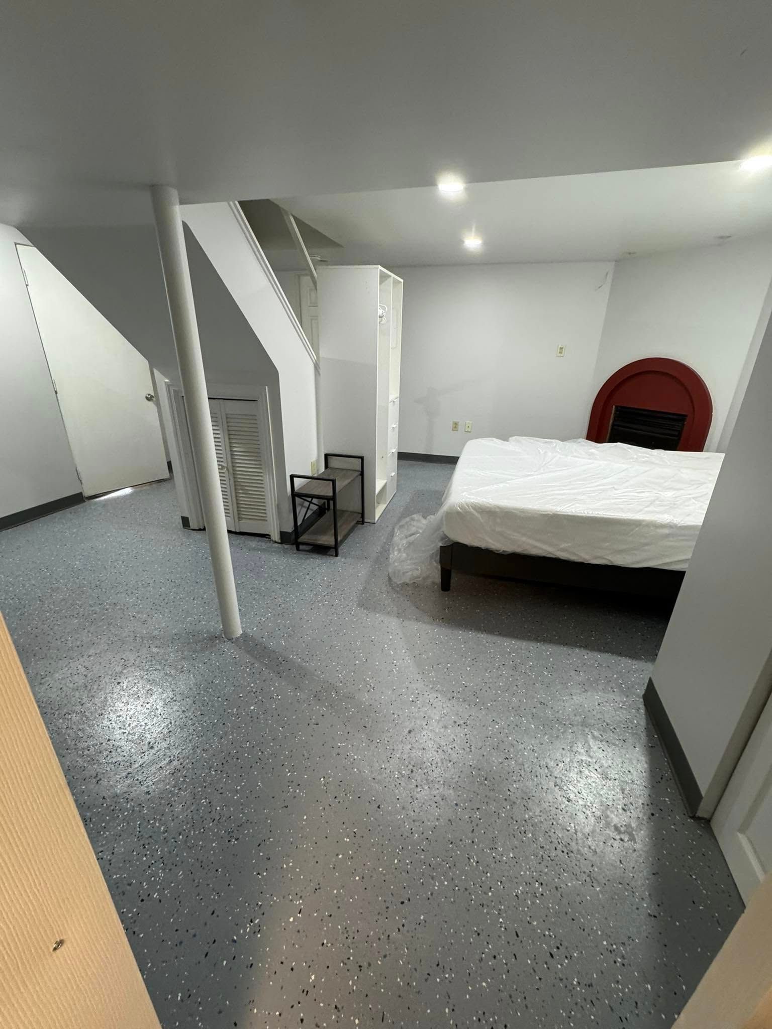 Basement bedroom with bed, fireplace, stairs, and gray speckled floor.