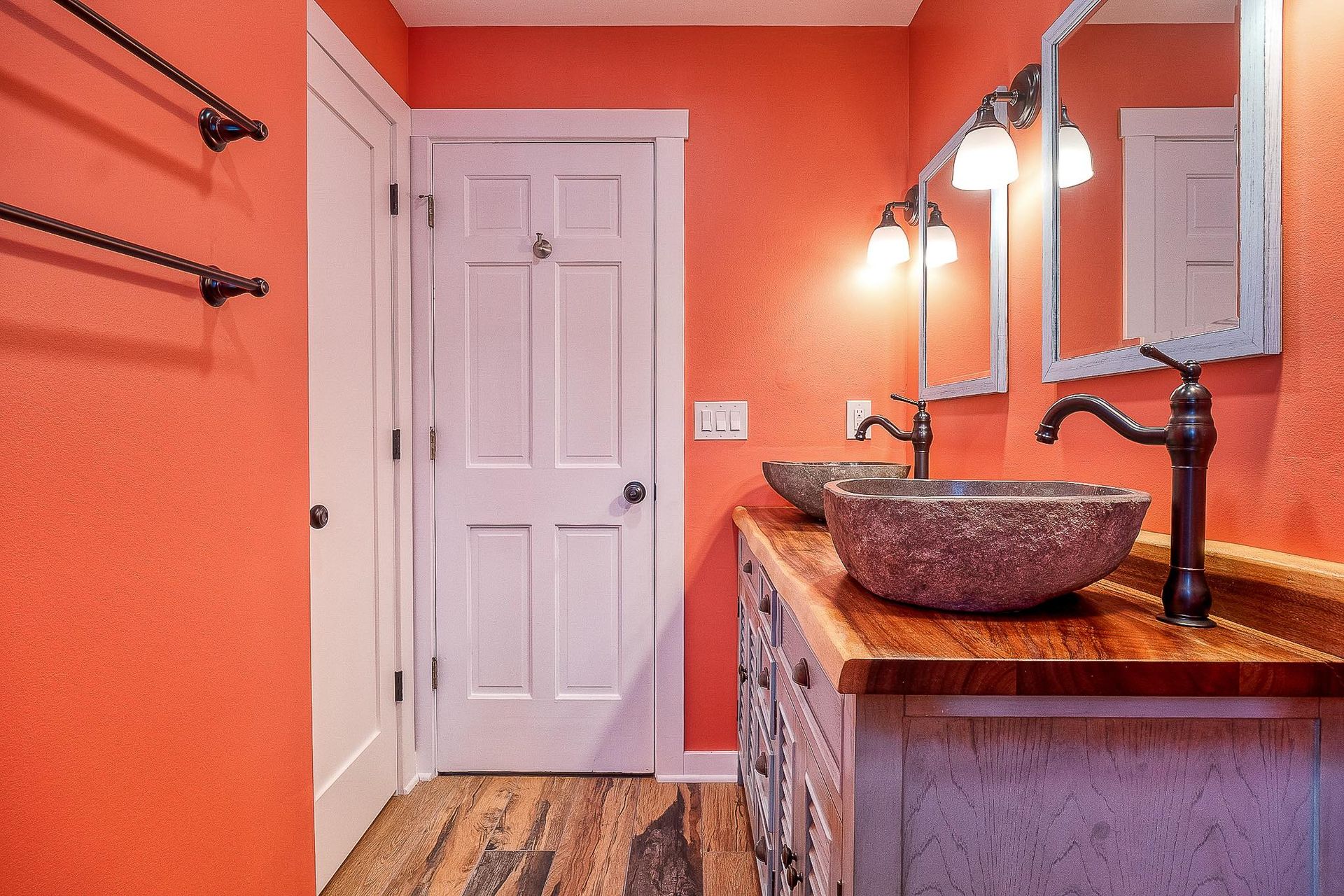 Coral-colored bathroom with double sinks and a wooden countertop. Two mirrors with sconces, white door.