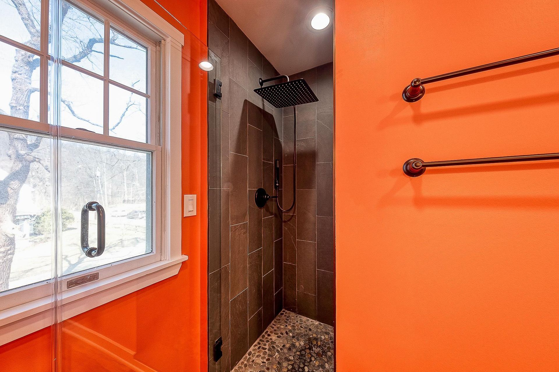 Orange-walled bathroom with shower. Dark tile inside, stone floor, window to left, two towel racks on the right.