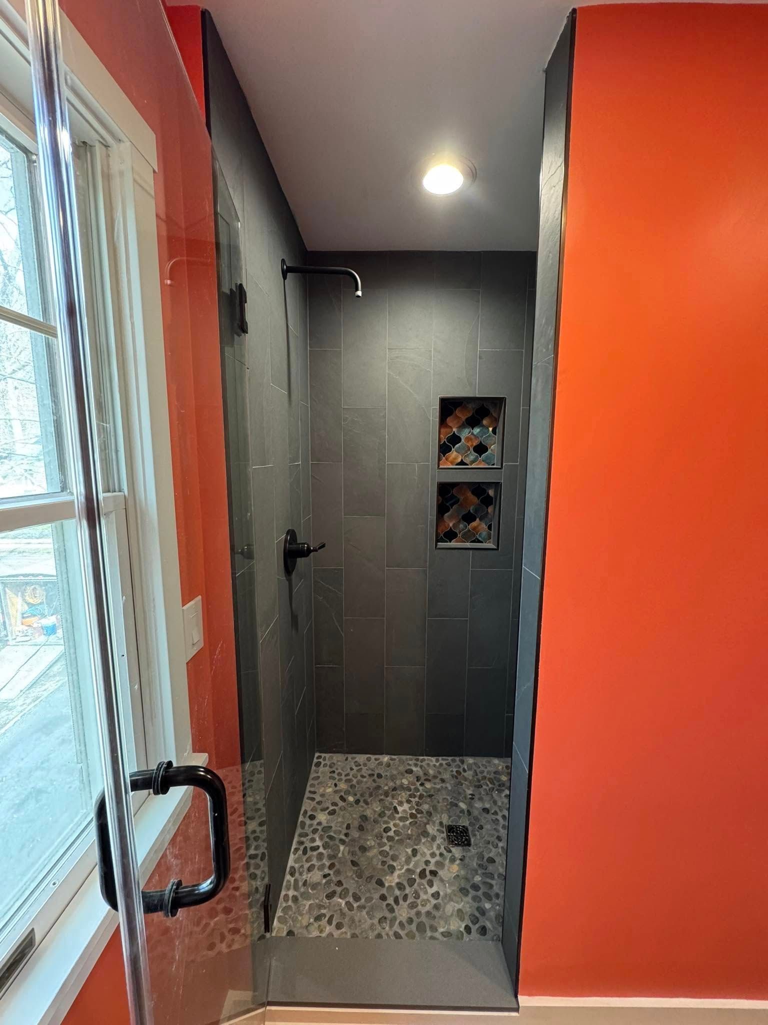 Narrow bathroom with gray tiled shower, pebble floor, built-in niches, and black fixtures; orange walls.