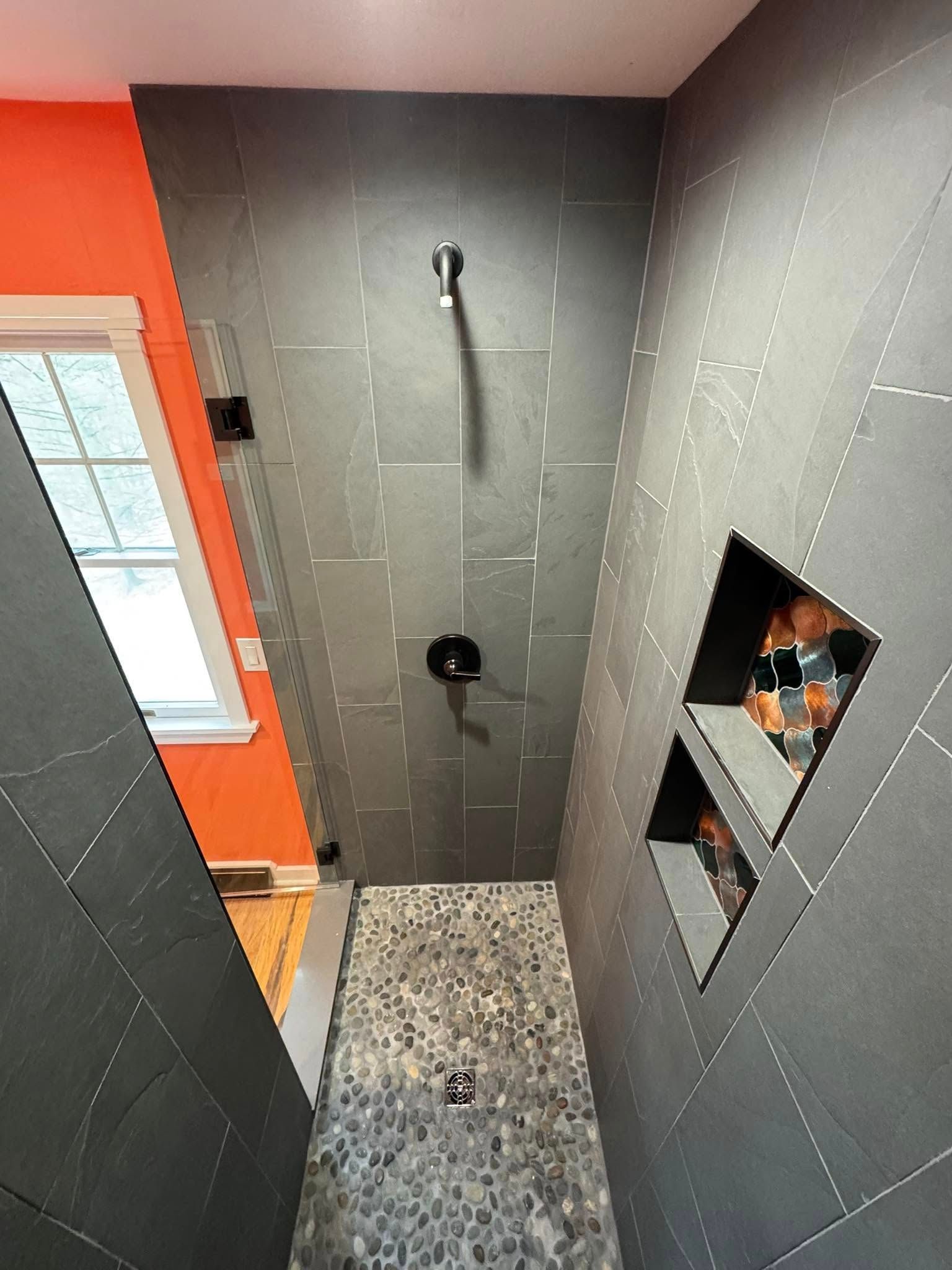 Modern shower with gray tile walls, pebble floor, and orange wall visible. Black shower fixtures and built-in niches.
