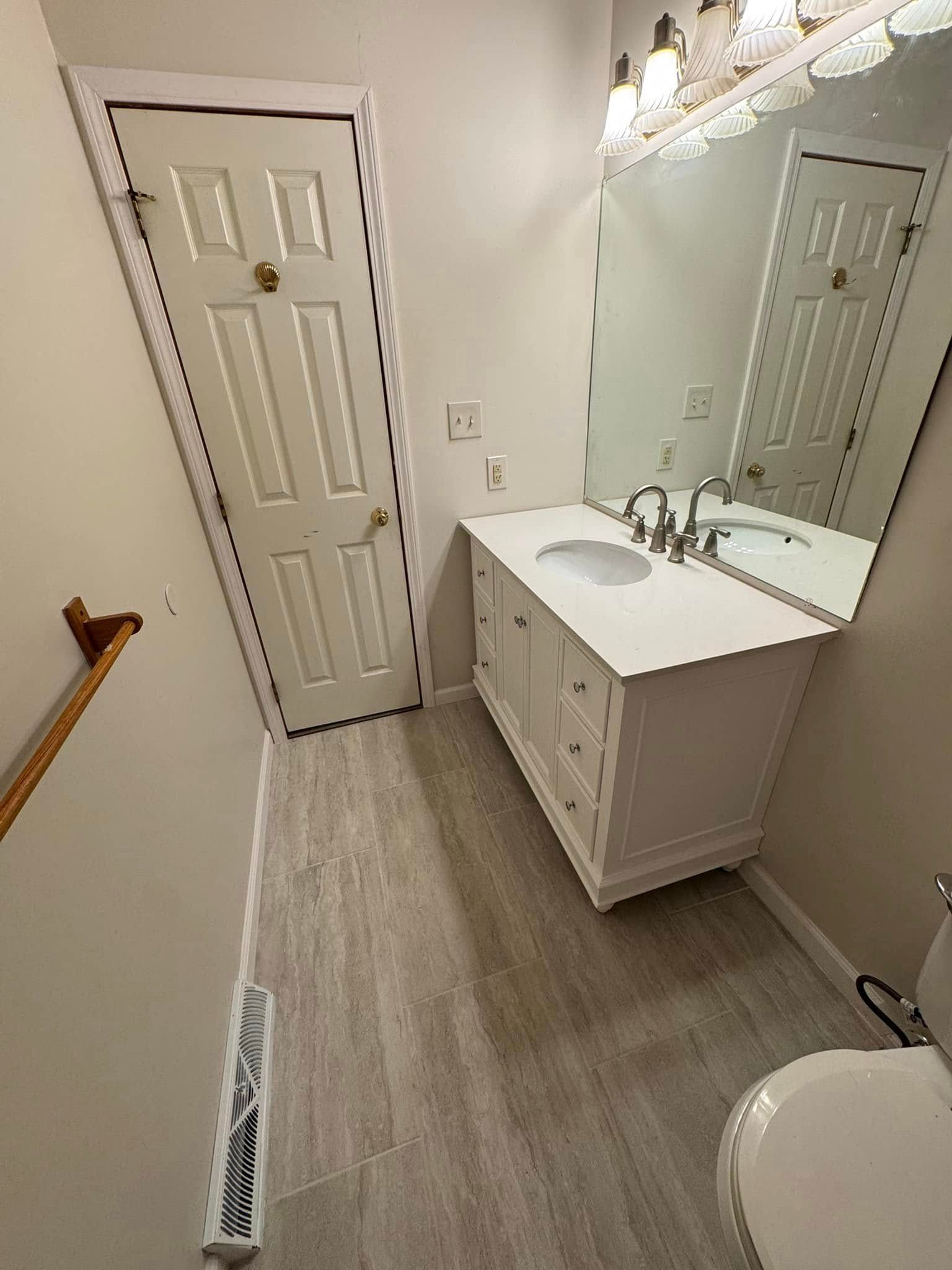 White bathroom with vanity, mirror, and toilet. Light wood-look flooring and a closed door are also visible.
