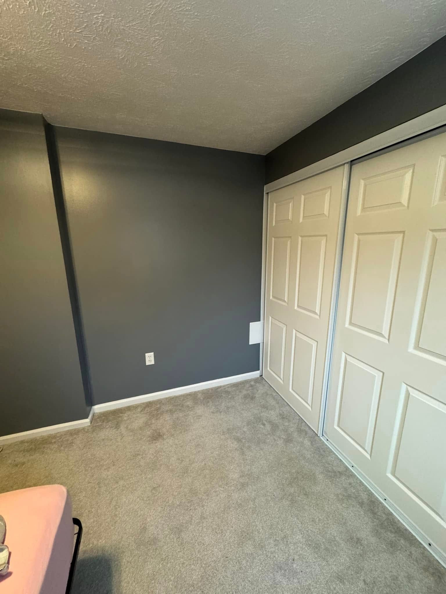 Gray-painted room with white closet doors and light-colored carpet. A pink bed frame is visible in the corner.