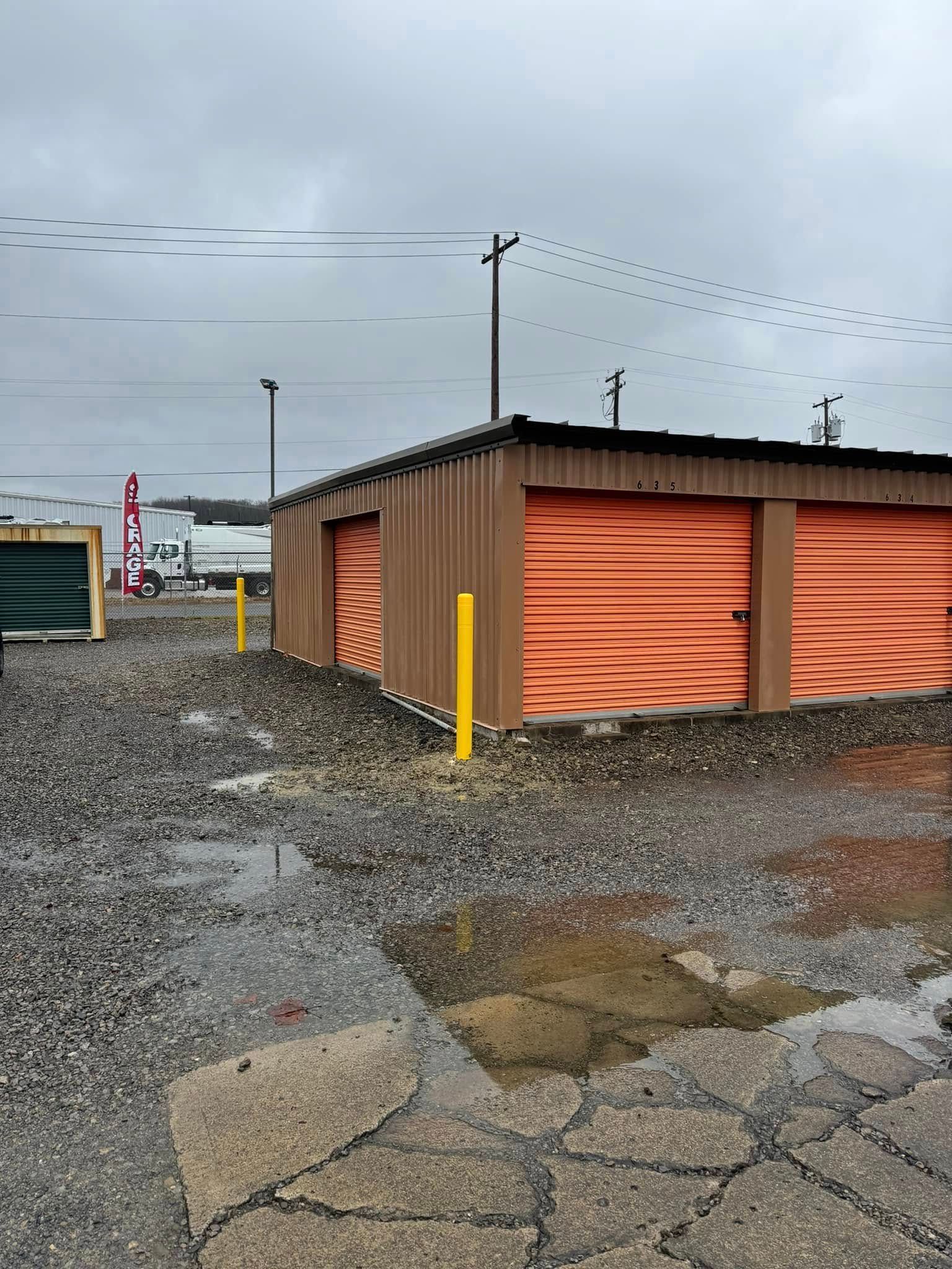 Storage units with orange doors, brown frames, and a gravel lot on an overcast day.