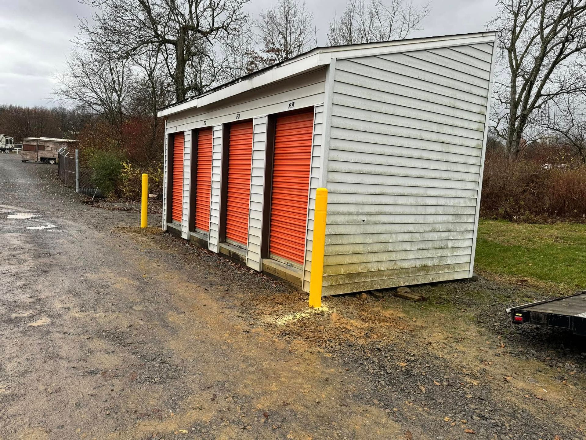 Storage units with orange doors, white siding, and yellow posts on a gravel drive.