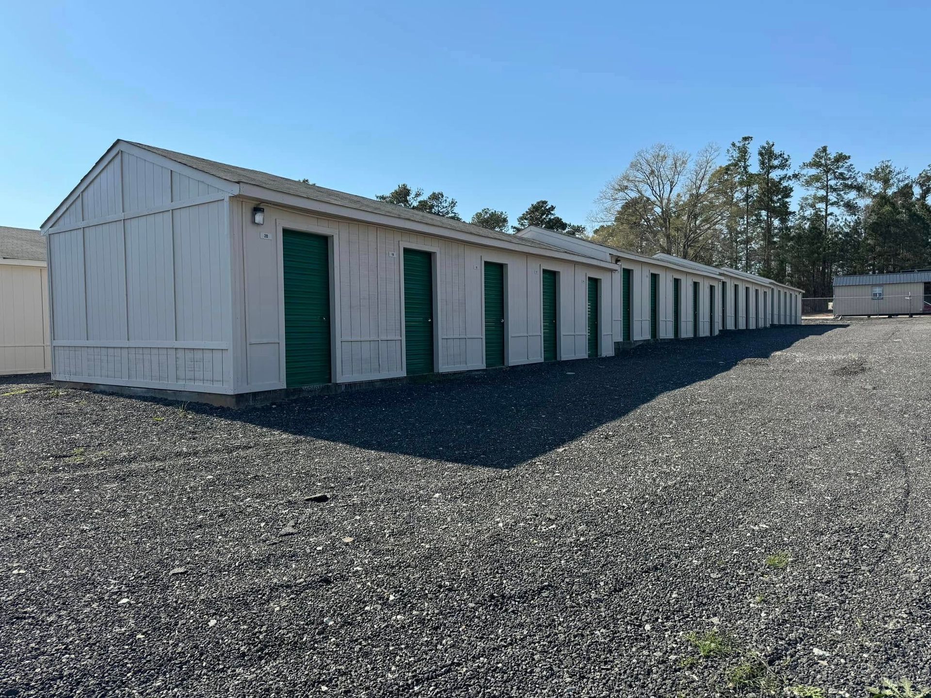 Row of white storage units with green doors, set on gravel, under a blue sky.
