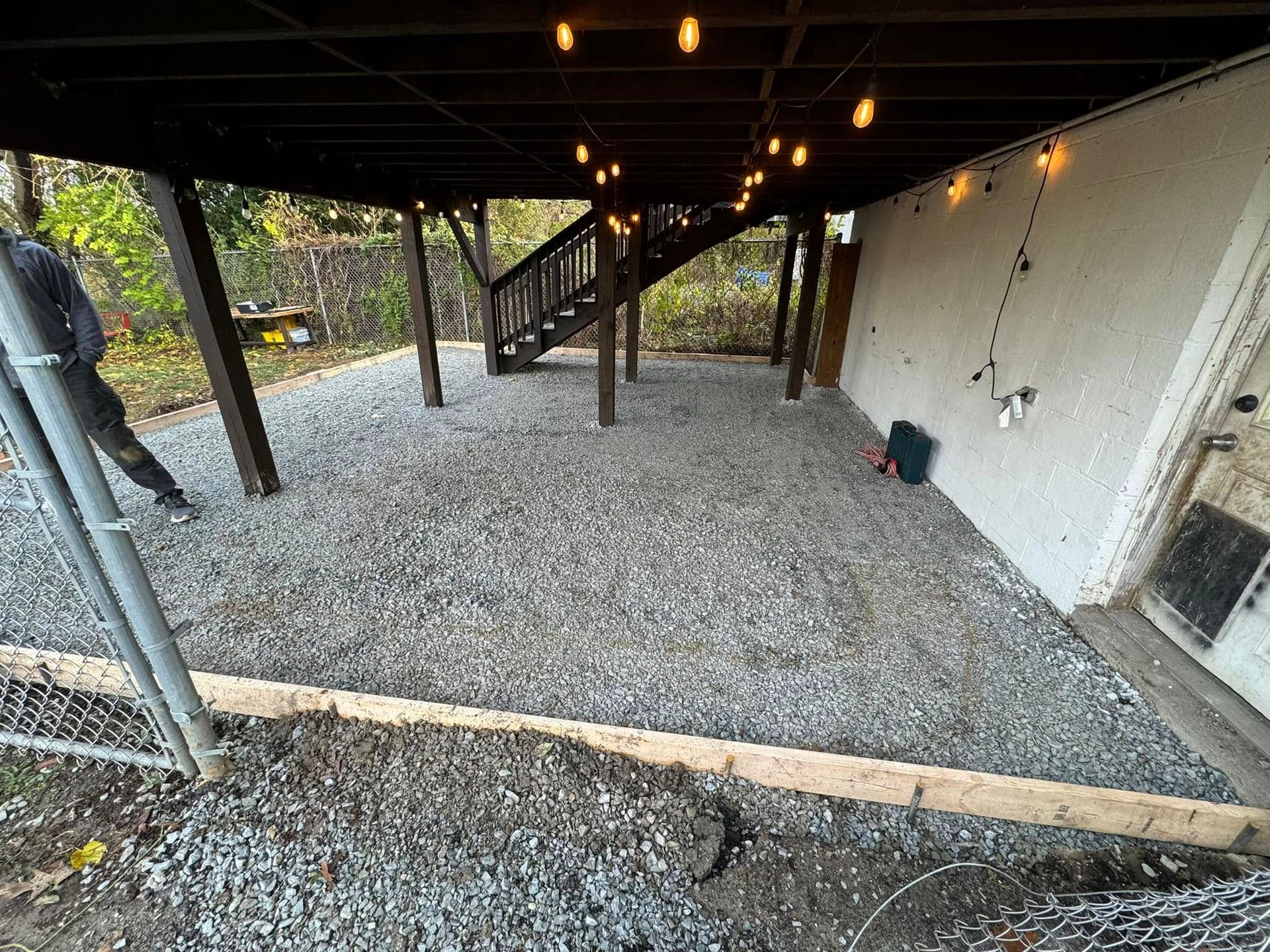 Gravel patio under a wooden deck with string lights. A person stands near a chain-link fence.