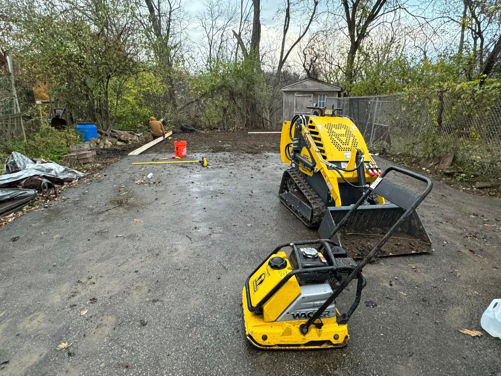 Construction scene: yellow skid steer and plate compactor on asphalt. Work area with trees and fence in background.