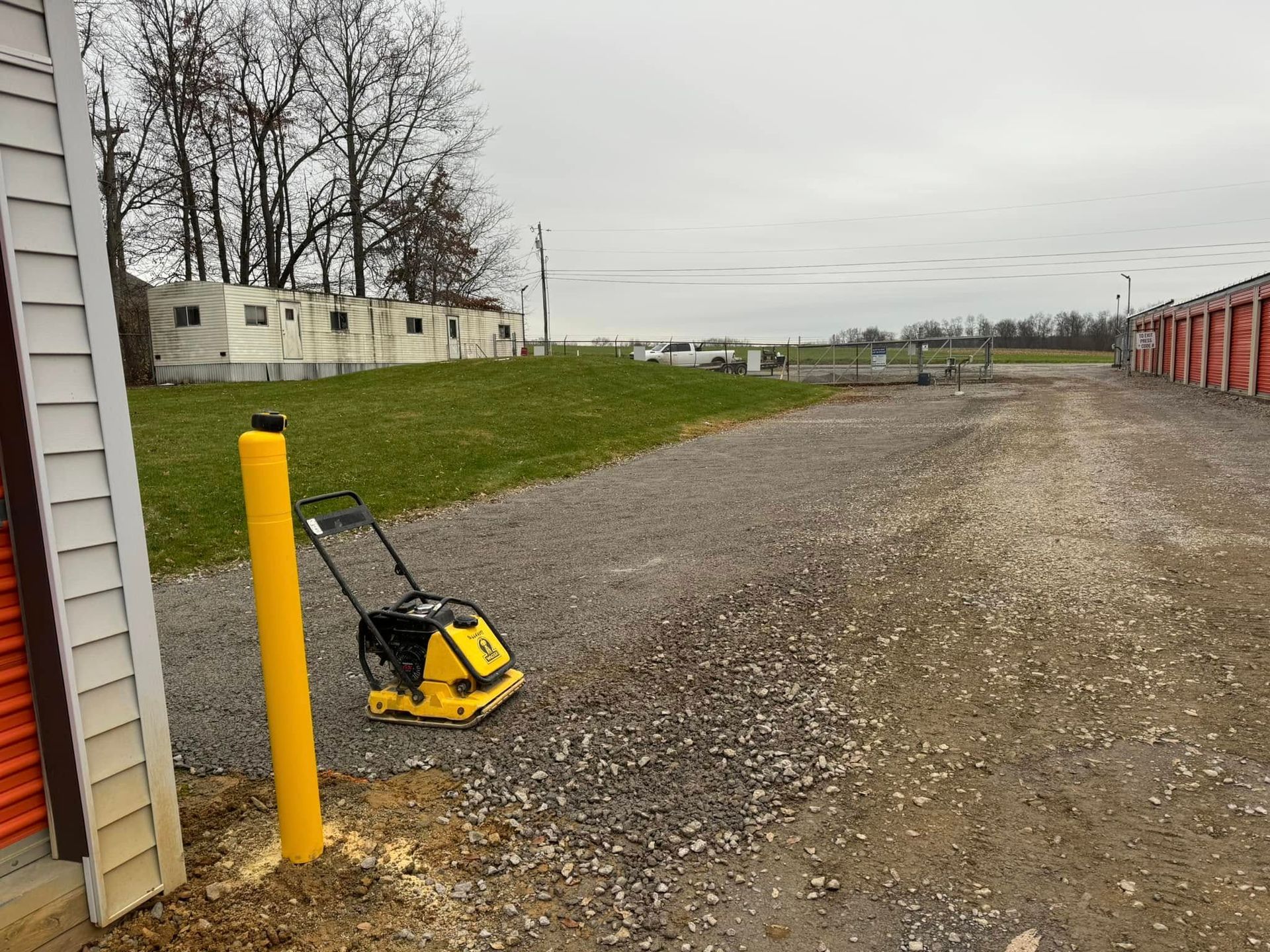 Gravel driveway with a yellow compactor, a yellow post, and storage units. Overcast sky.
