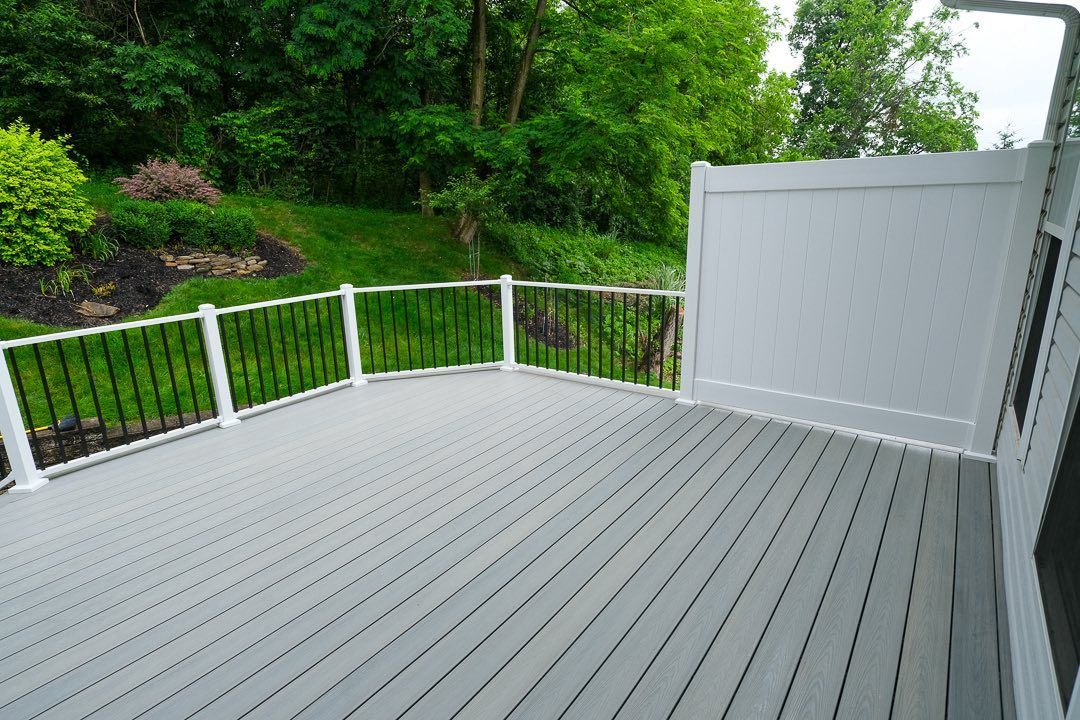 Gray composite deck with white and black railing, adjacent to a white privacy fence. Green foliage in the background.