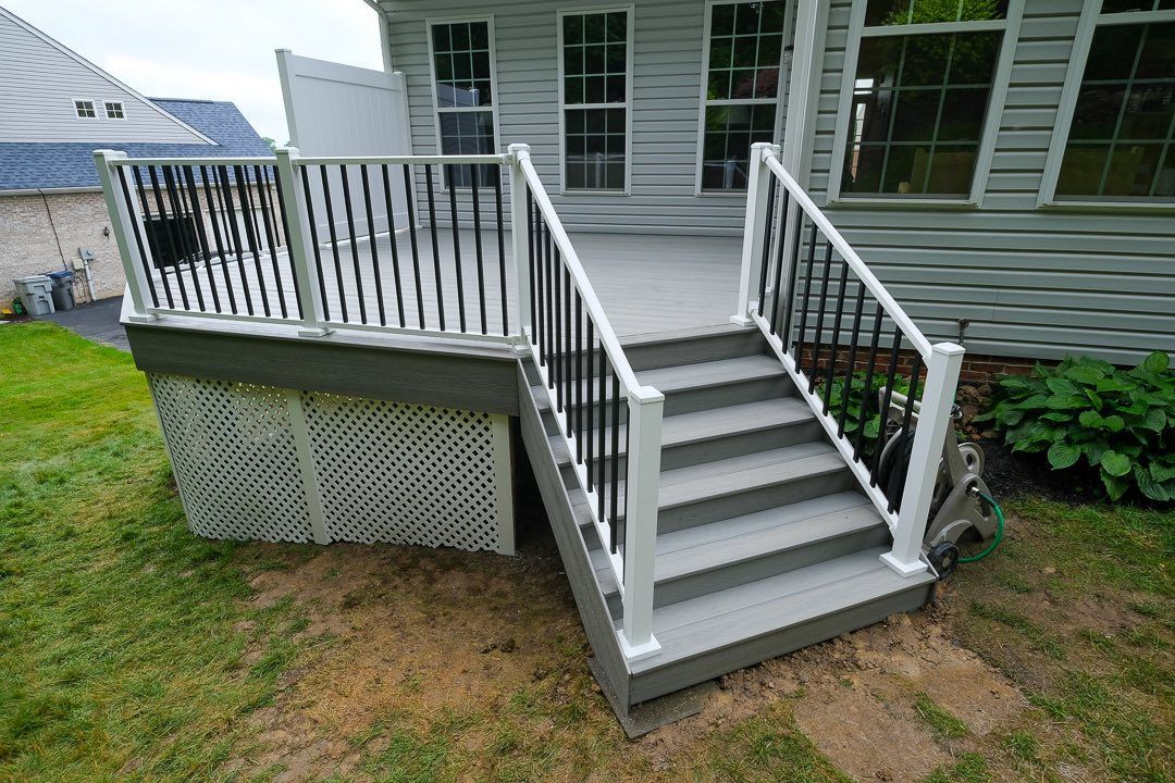 Gray deck with stairs and black and white railing, near a house with windows.