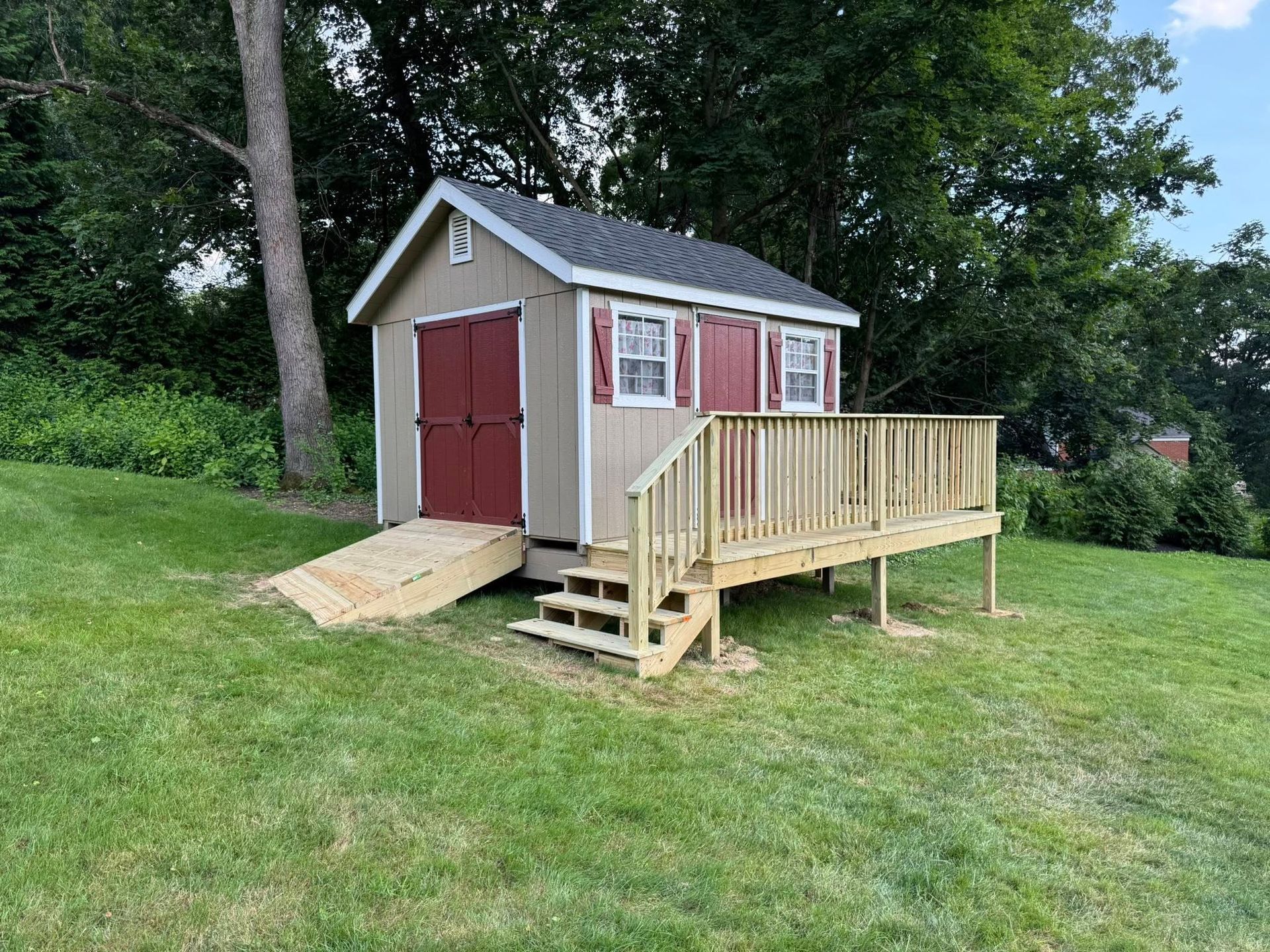 Tan shed with red doors, deck, and ramp on a grassy lawn.