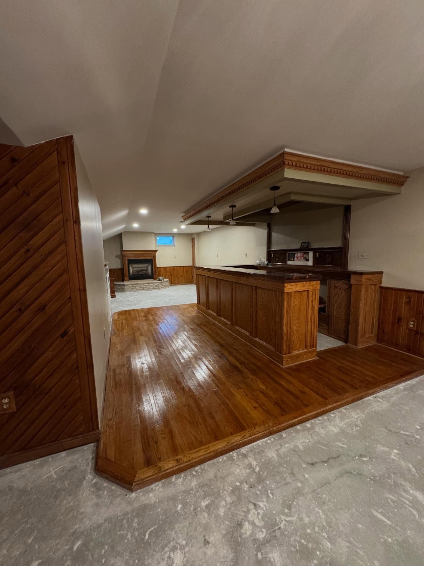 Basement bar with wood paneling, wood floor, and fireplace. Empty, with bar and overhead lighting.