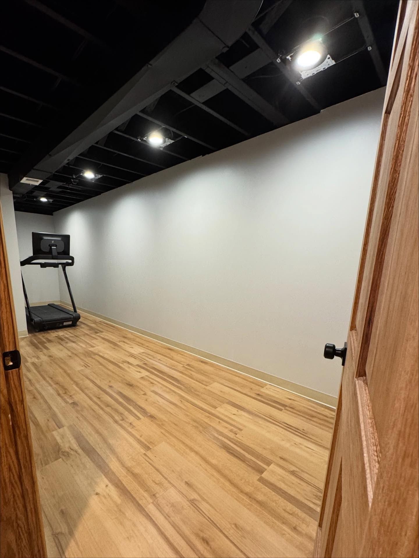 Basement gym with a treadmill, hardwood floor, white wall, black ceiling, and open wooden door.