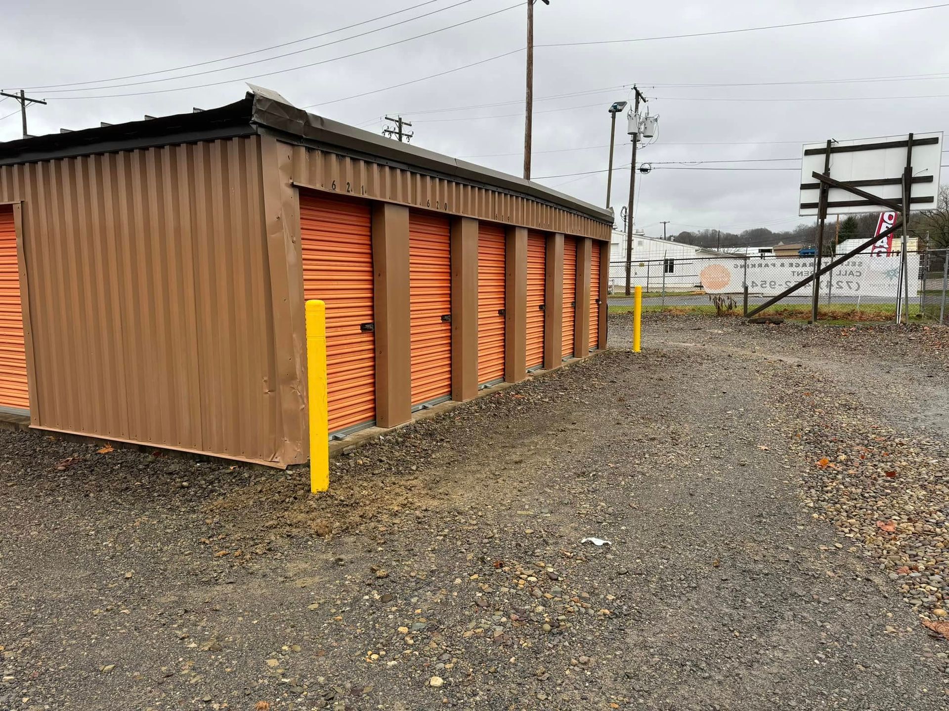 Brown and orange storage units with yellow posts in a gravel lot under a cloudy sky.