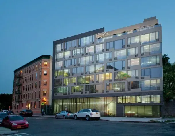 Modern glass-front apartment building at dusk beside an older brick building, with parked cars out front