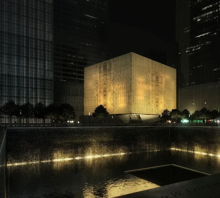 Golden-lit cube memorial reflected in a dark water pool at night, surrounded by tall buildings.