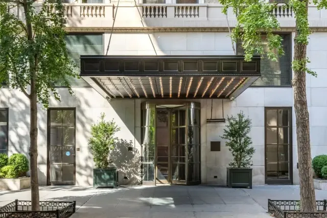 Modern building entrance with glass doors, white facade, balconies, and trees lining the sidewalk.